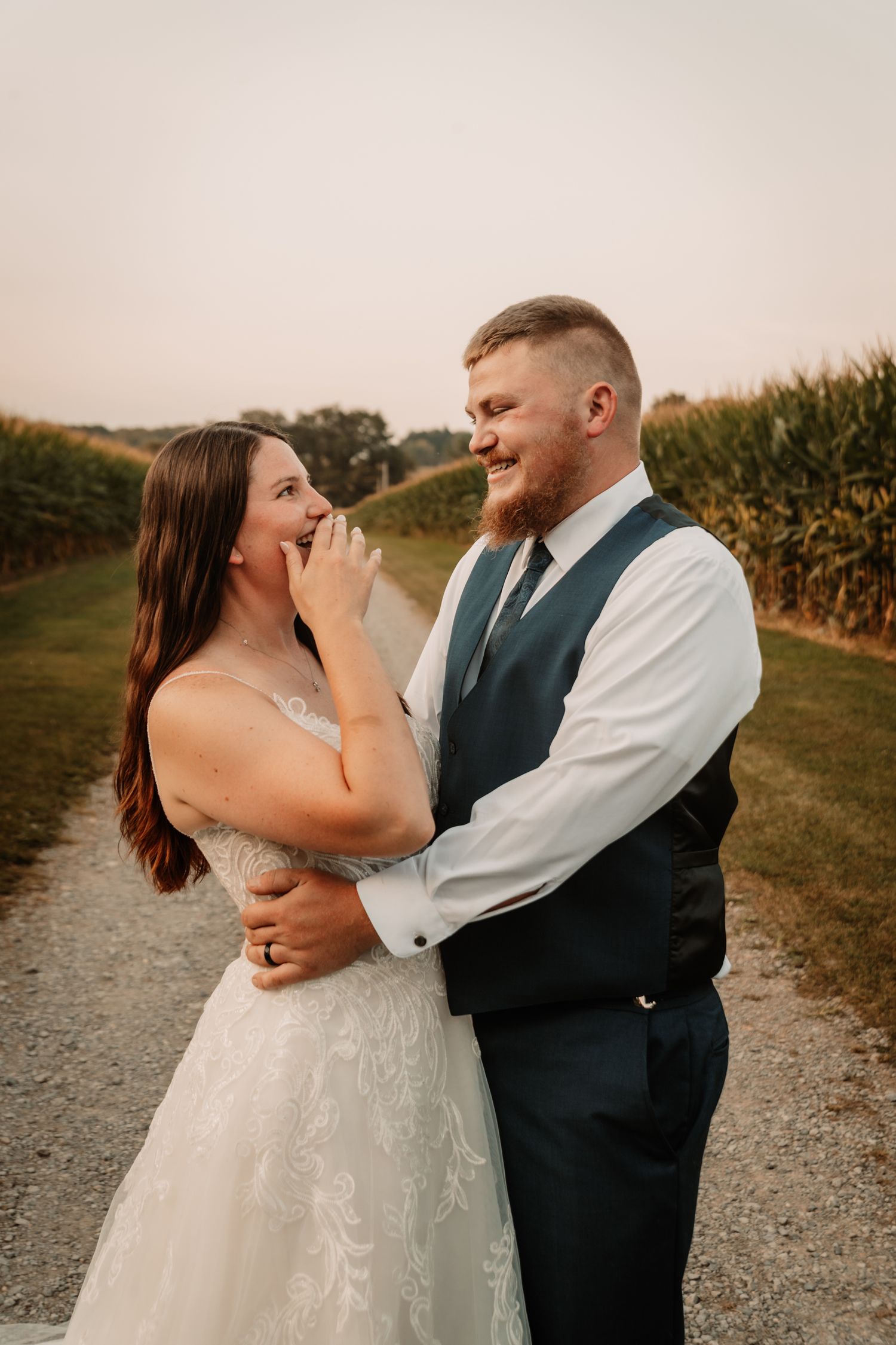 Couple shares joyful candid moment together on rural path surrounded by cornfields at sunset.