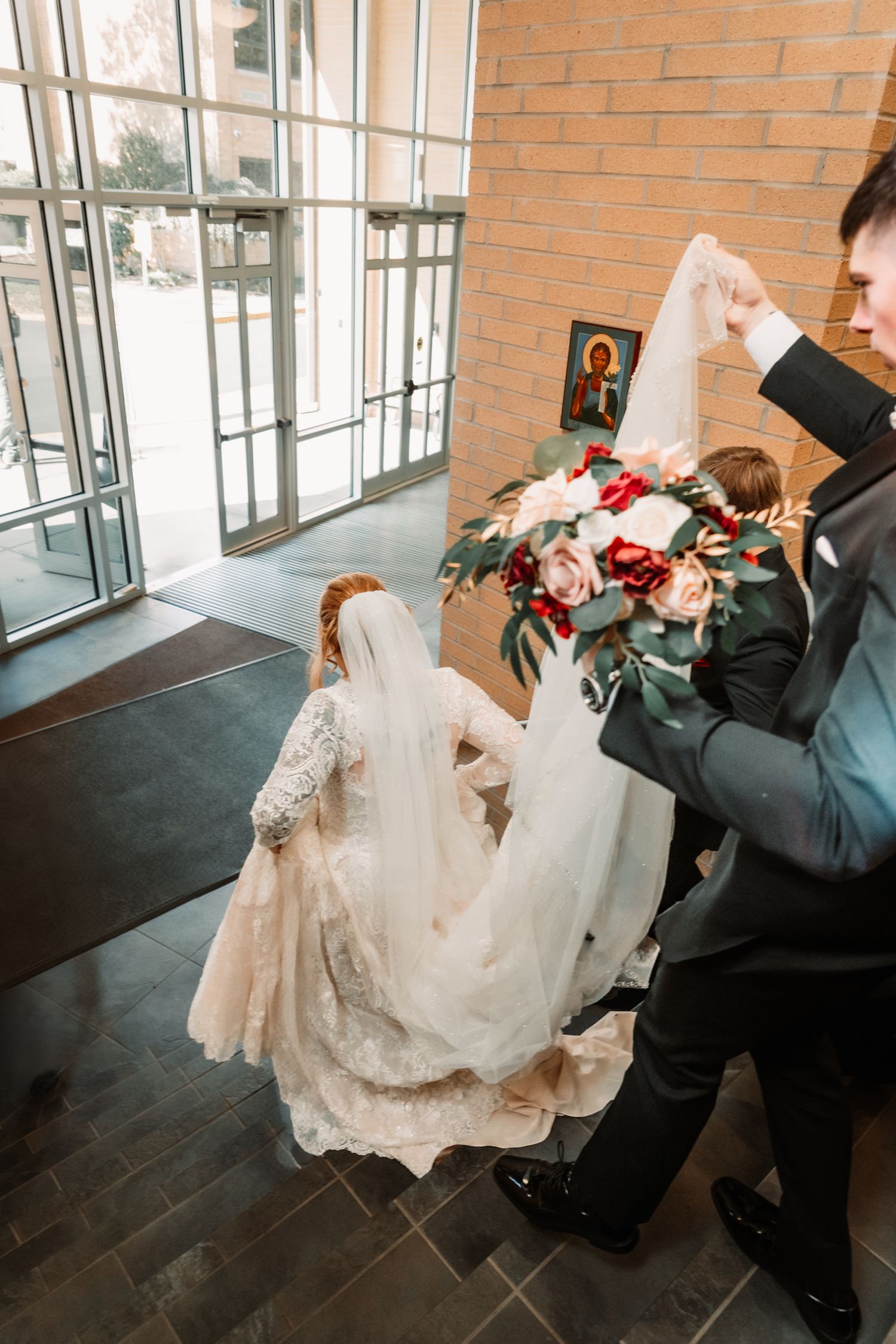 A wedding dress with lace details and flowing veil lies draped beside a bouquet of pink and red roses near glass doors.
