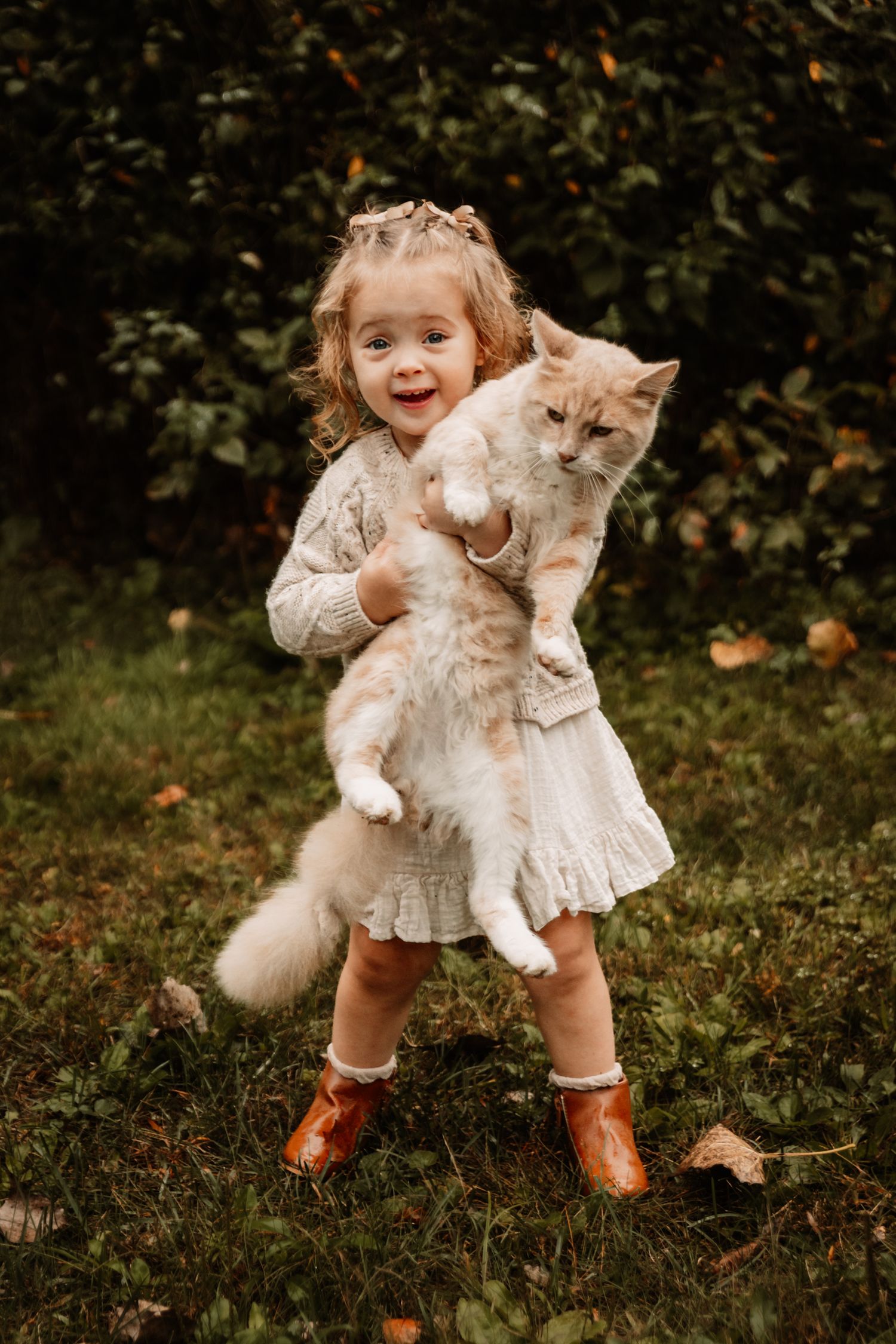A child in a white dress and brown boots holds a fluffy cream-colored cat outdoors during autumn.