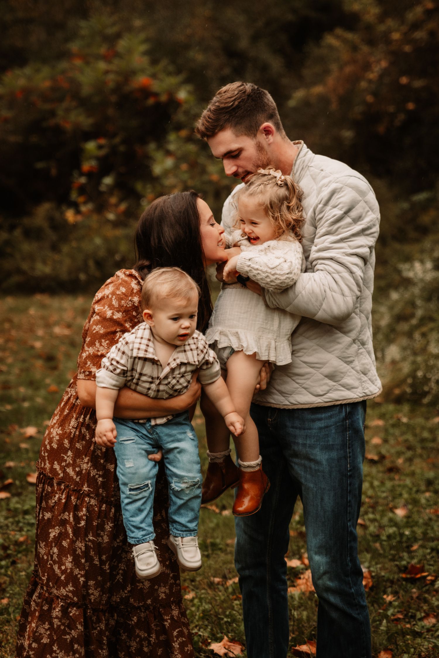 A family shares a tender autumn moment outdoors with their two young children among fallen leaves.