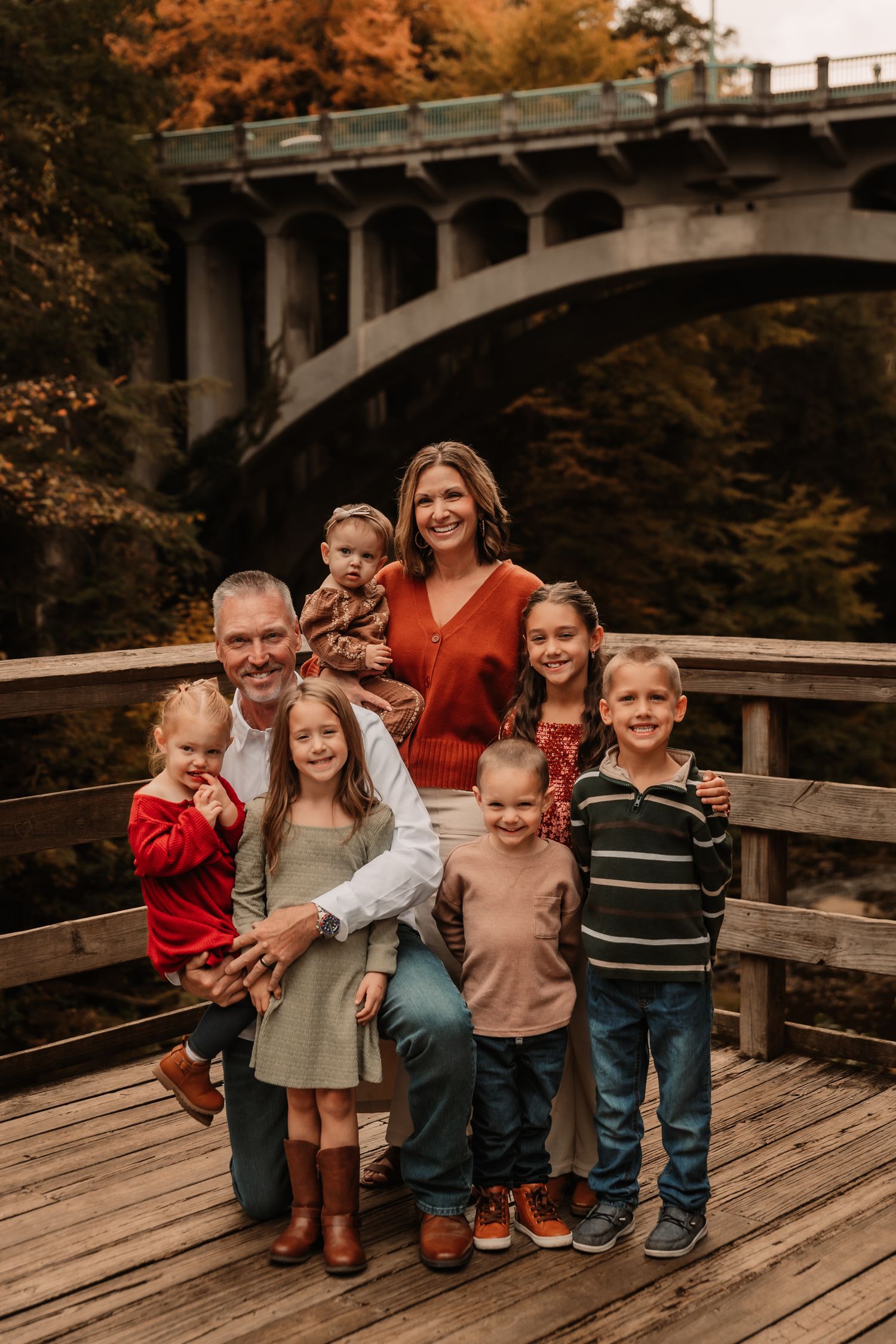 A family poses together on a wooden deck with a large stone bridge and autumn foliage in the background.