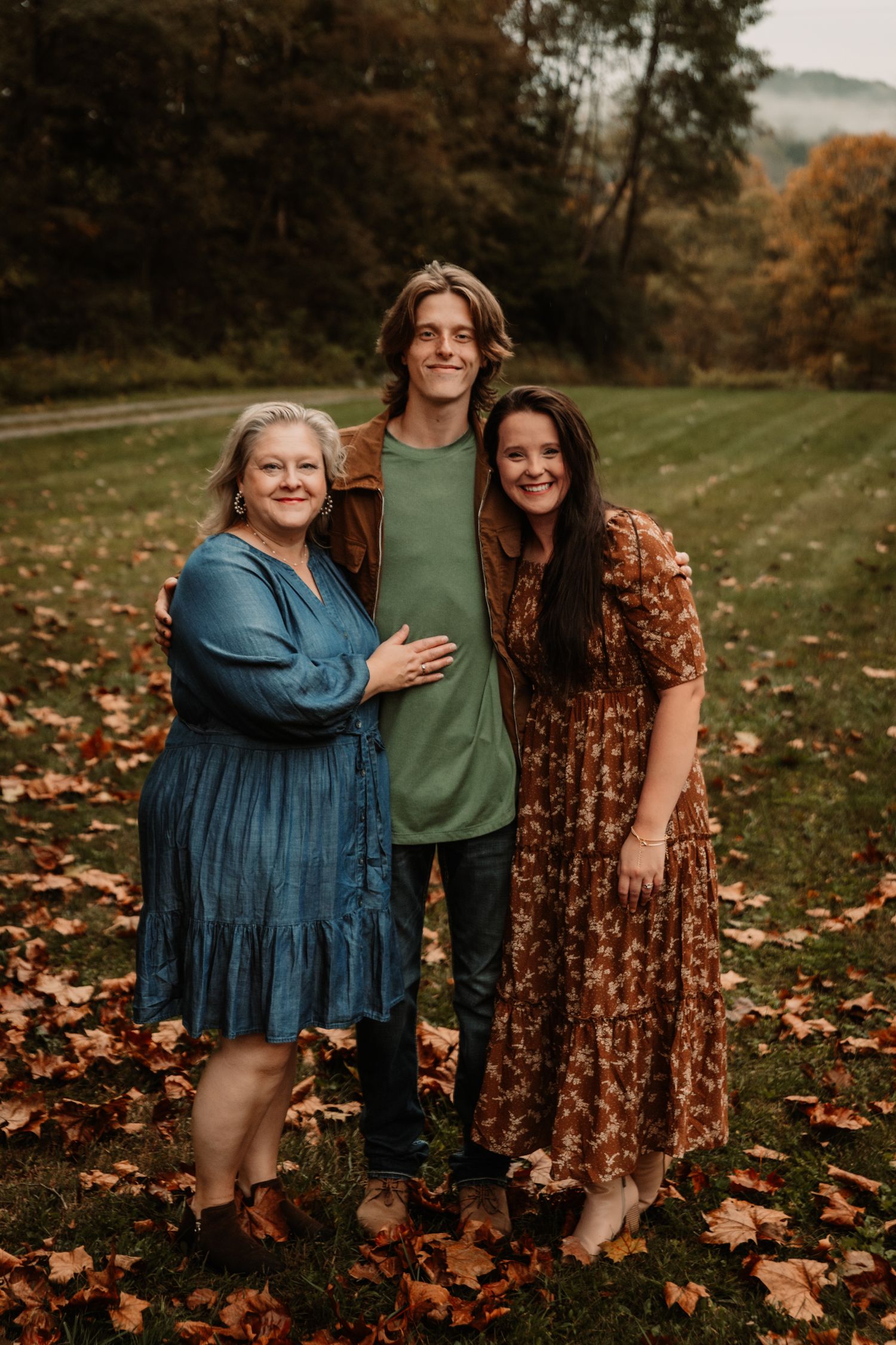 Three people pose together outdoors on an autumn day surrounded by fallen leaves.