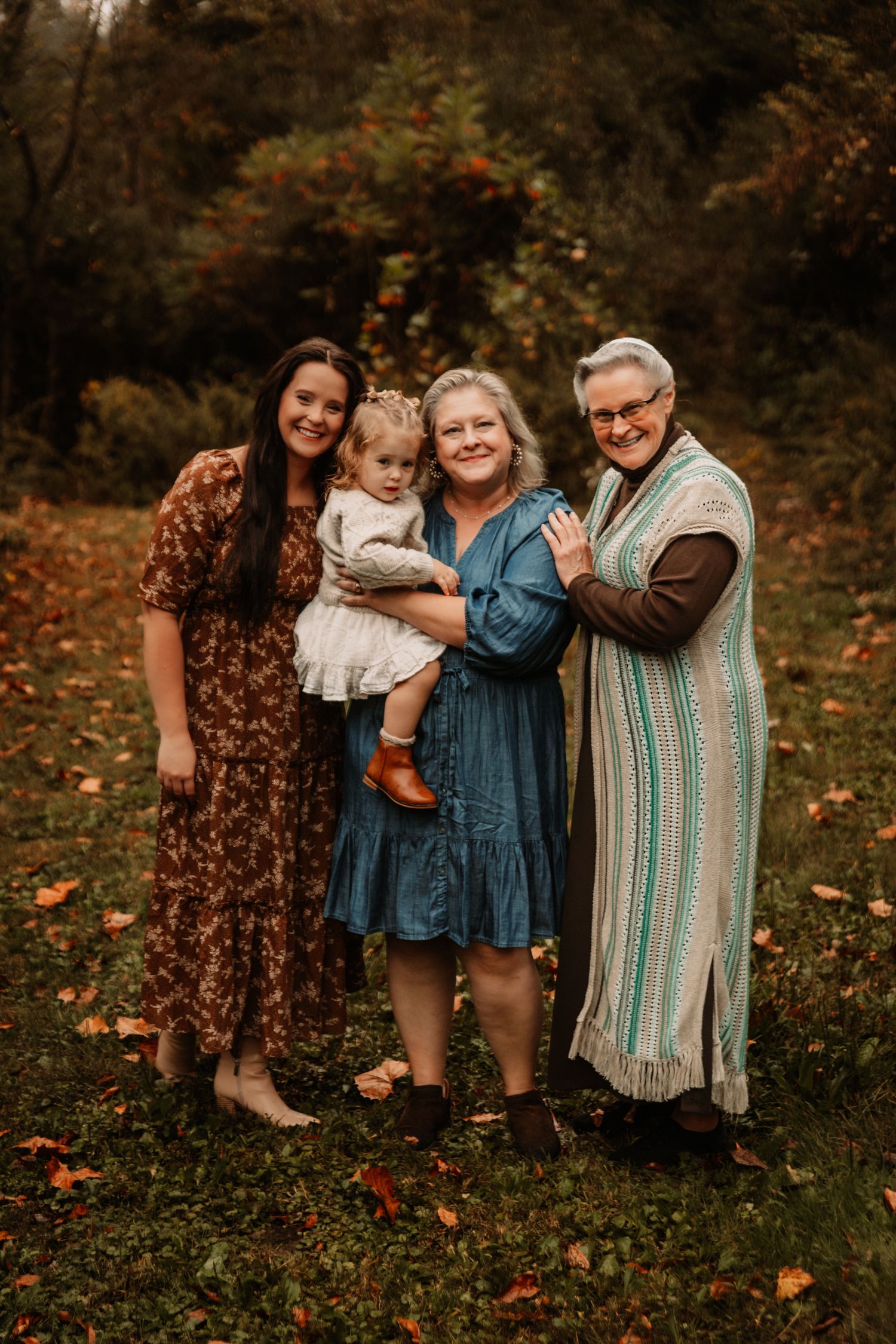 Four generations pose together in a wooded autumn setting wearing coordinating floral and striped dresses and sweaters.