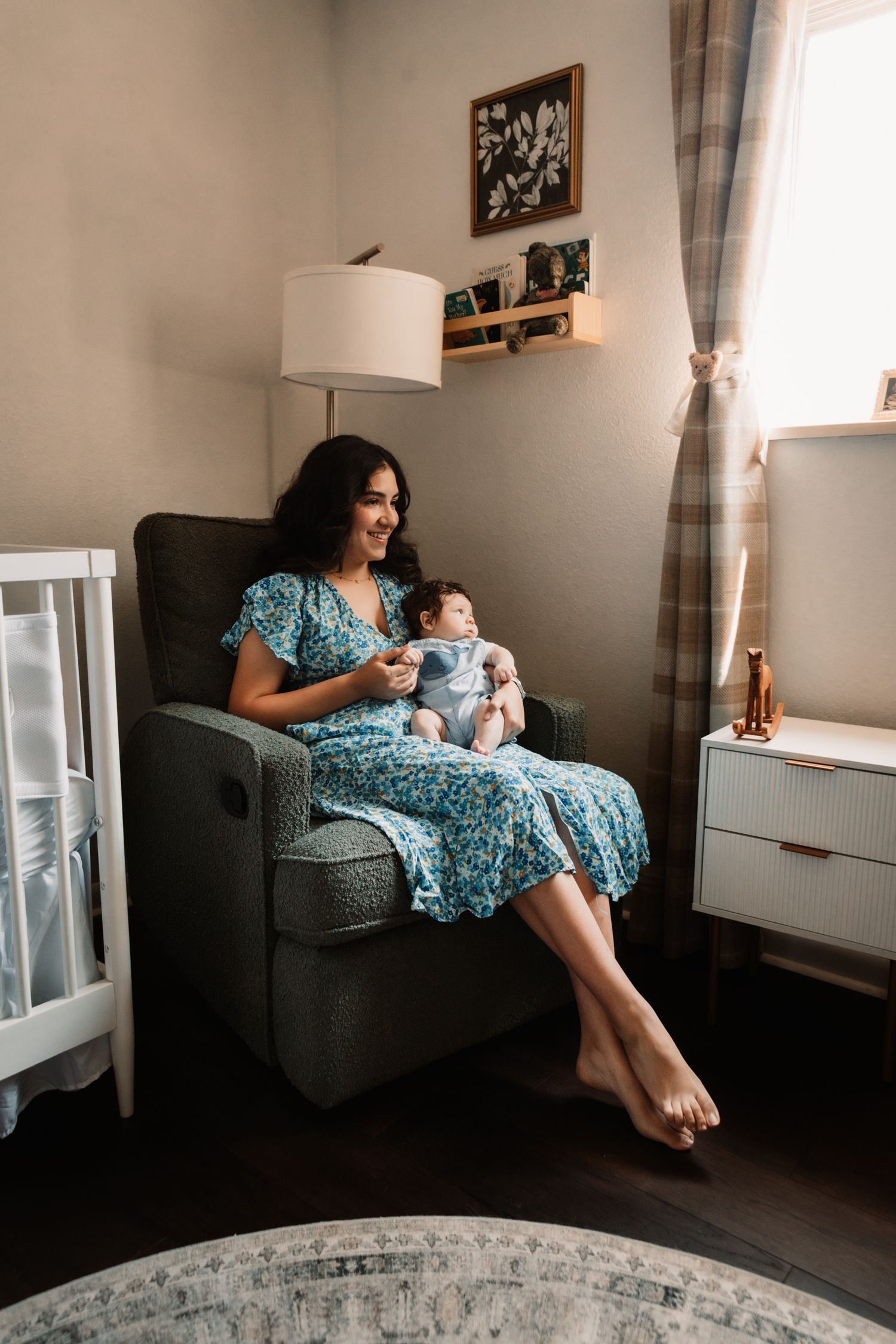 A peaceful nursery scene with someone in a blue floral dress relaxing in a gray armchair with an infant in a serene setting.