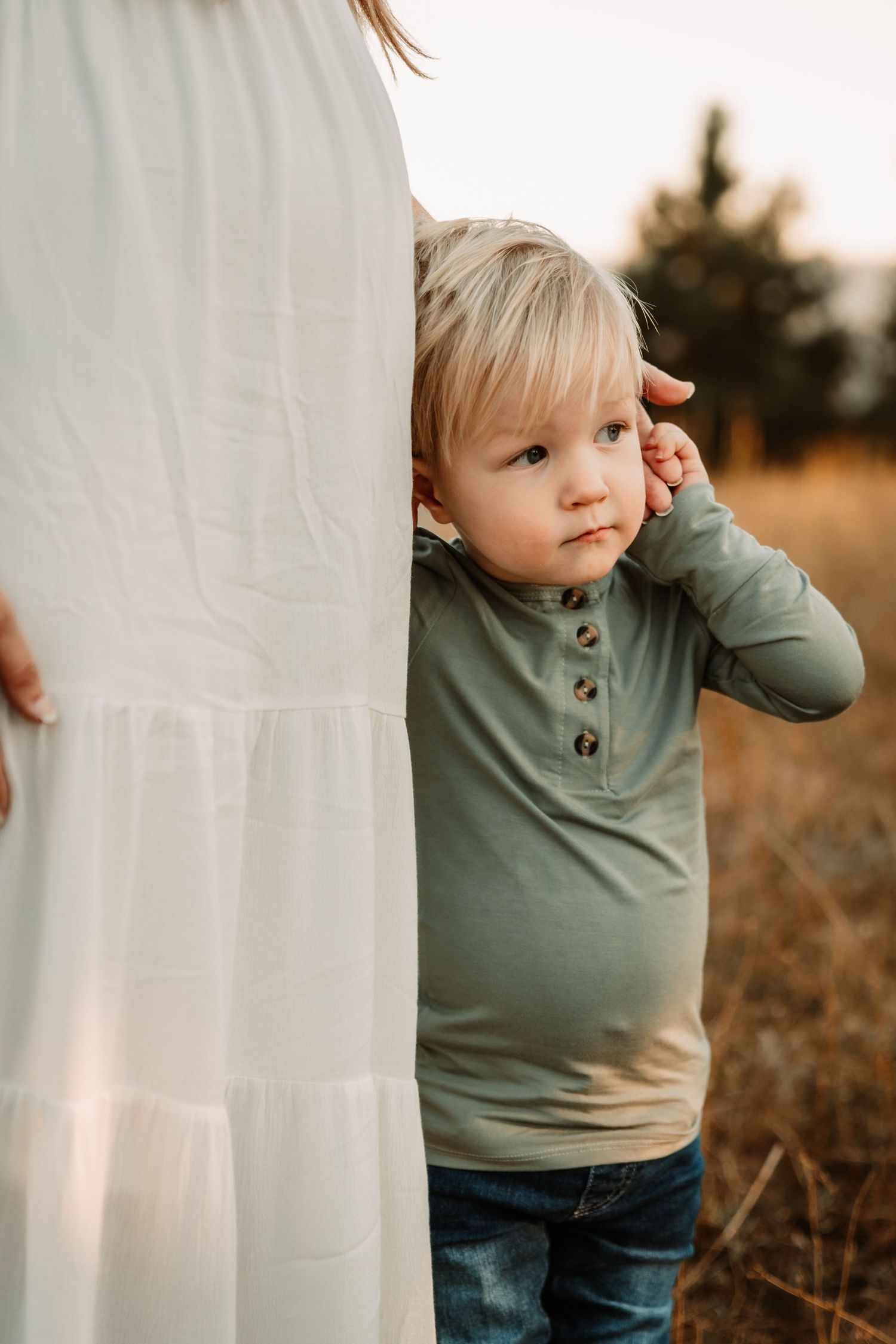 A small child in a sage green henley shirt peeks out from behind a white flowing dress in an outdoor setting at sunset.