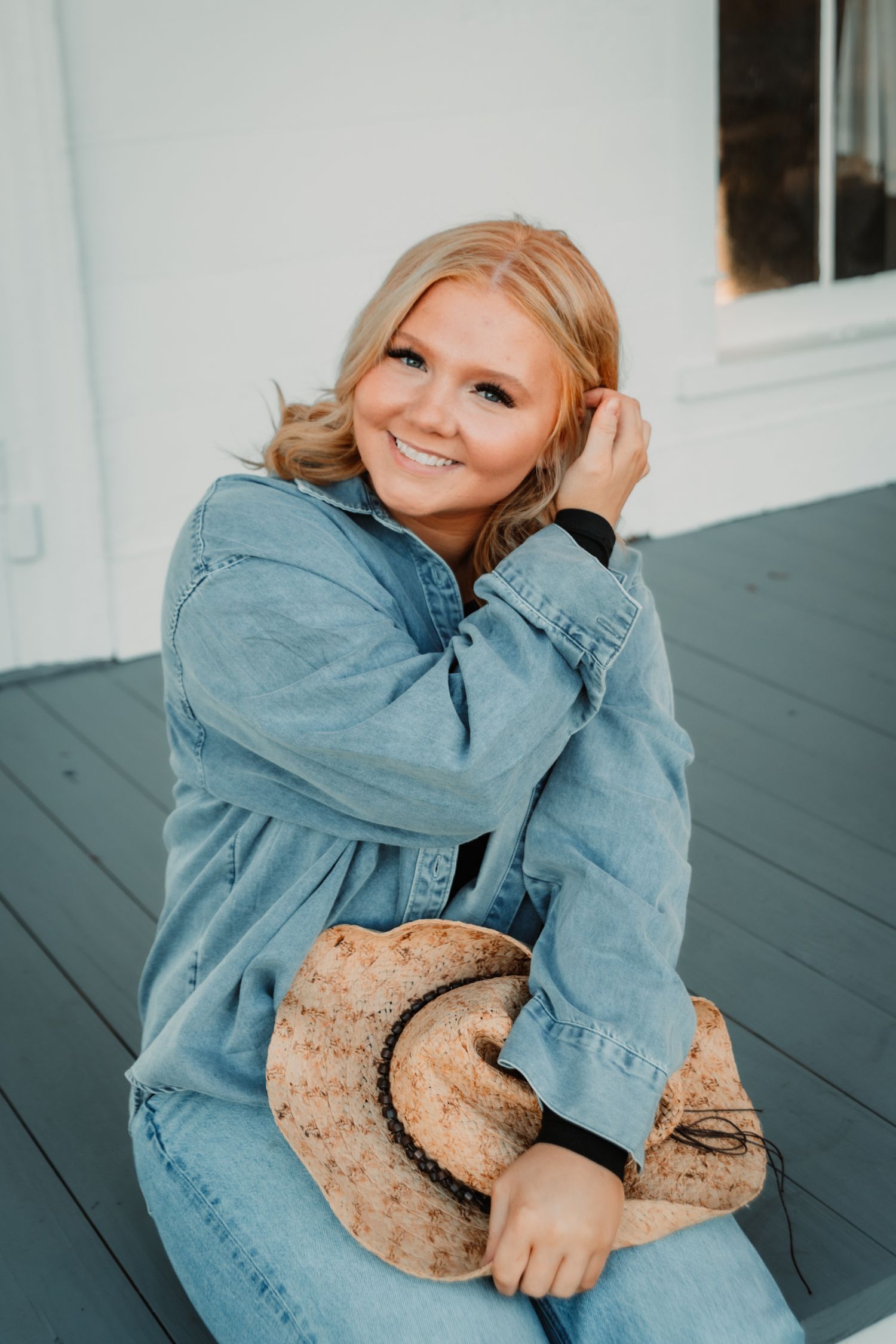 A person in denim jacket smiles while sitting on a dark porch and touching their blonde hair.