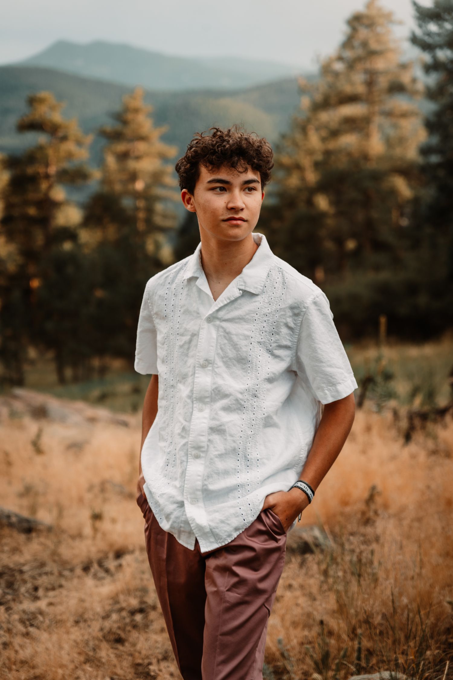Portrait of a young person in a white button-down shirt and brown pants standing against a mountain landscape backdrop.
