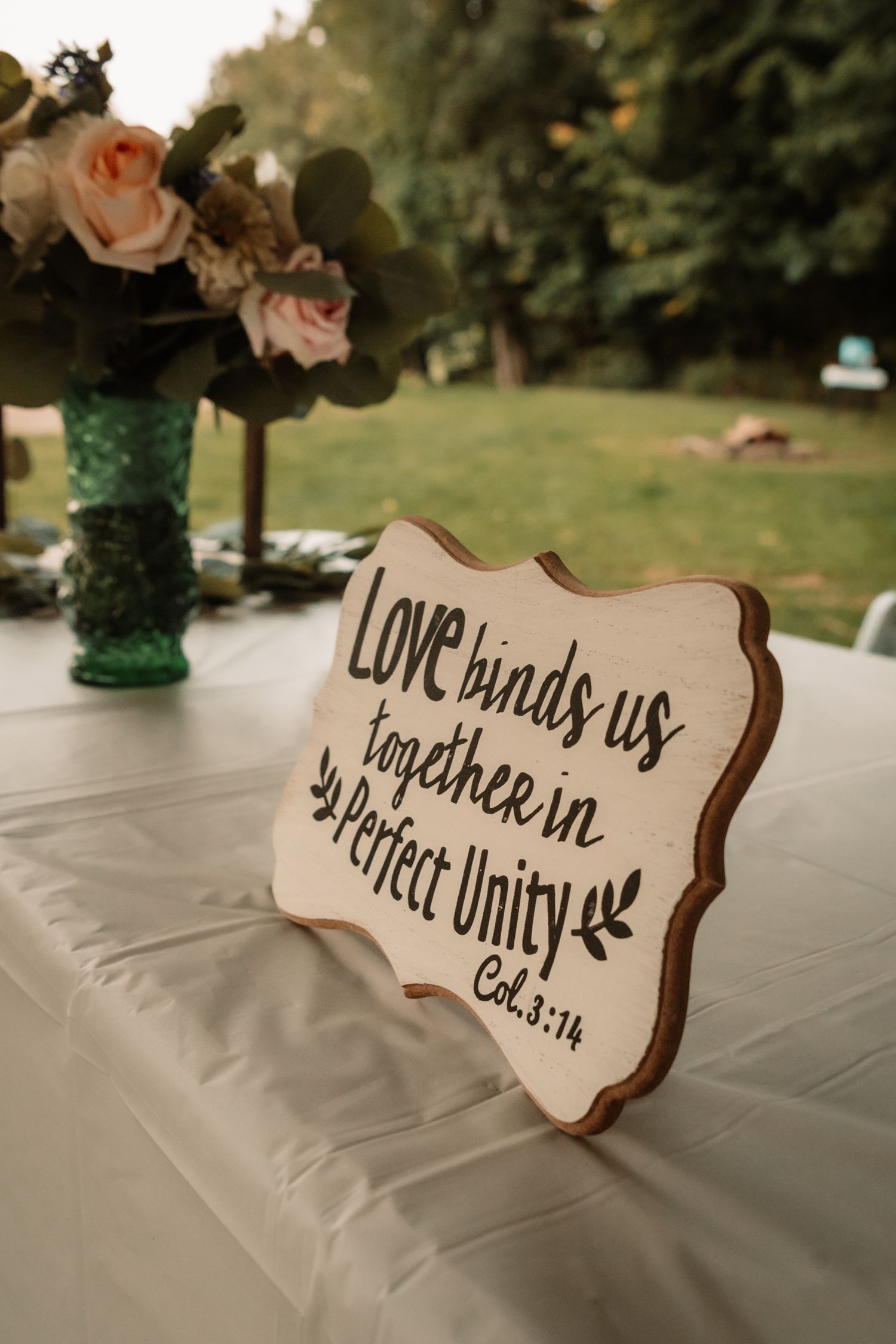 A wooden wedding sign reads 'Love binds us together in perfect unity' next to pink roses in a green vase on a white tablecloth.