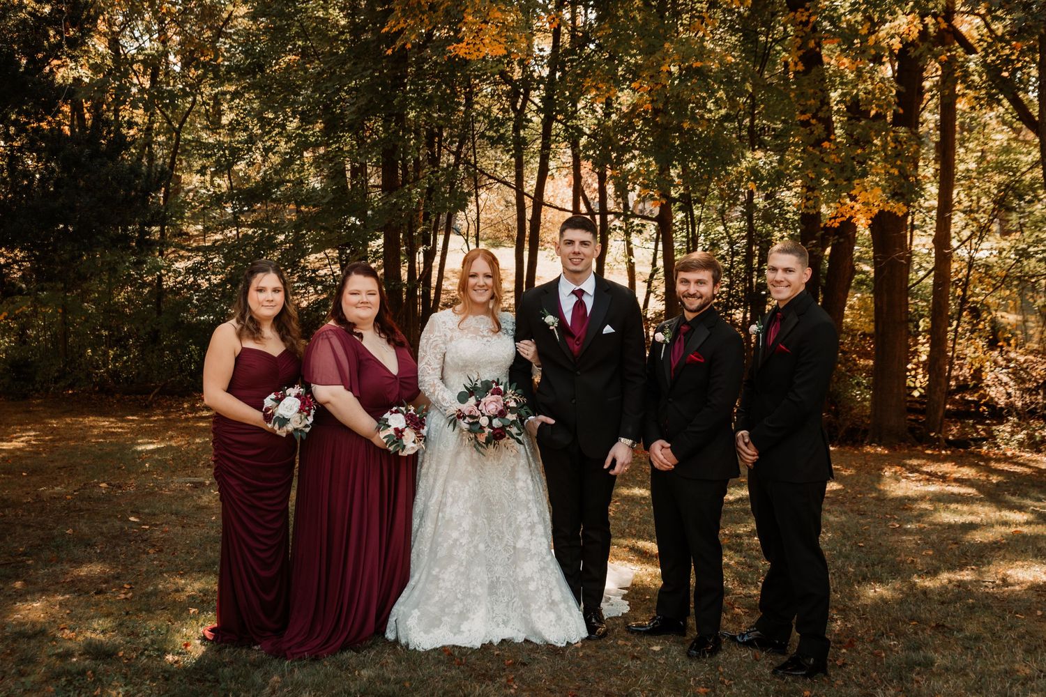 A wedding party poses in a sunlit autumn forest with burgundy bridesmaids dresses and formal black attire.
