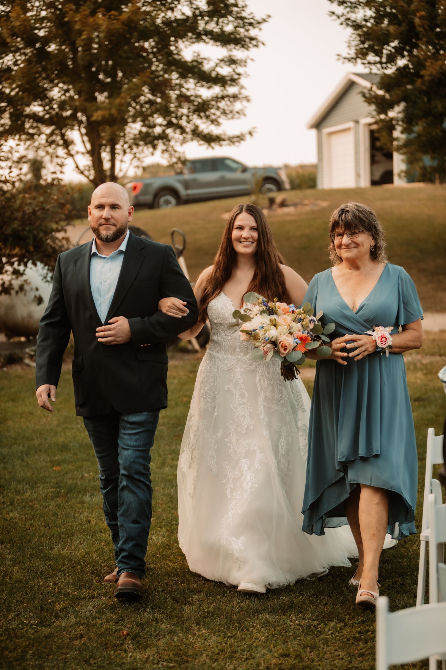 Bridal party walks down grassy path at outdoor wedding venue with car and white building in background at sunset.