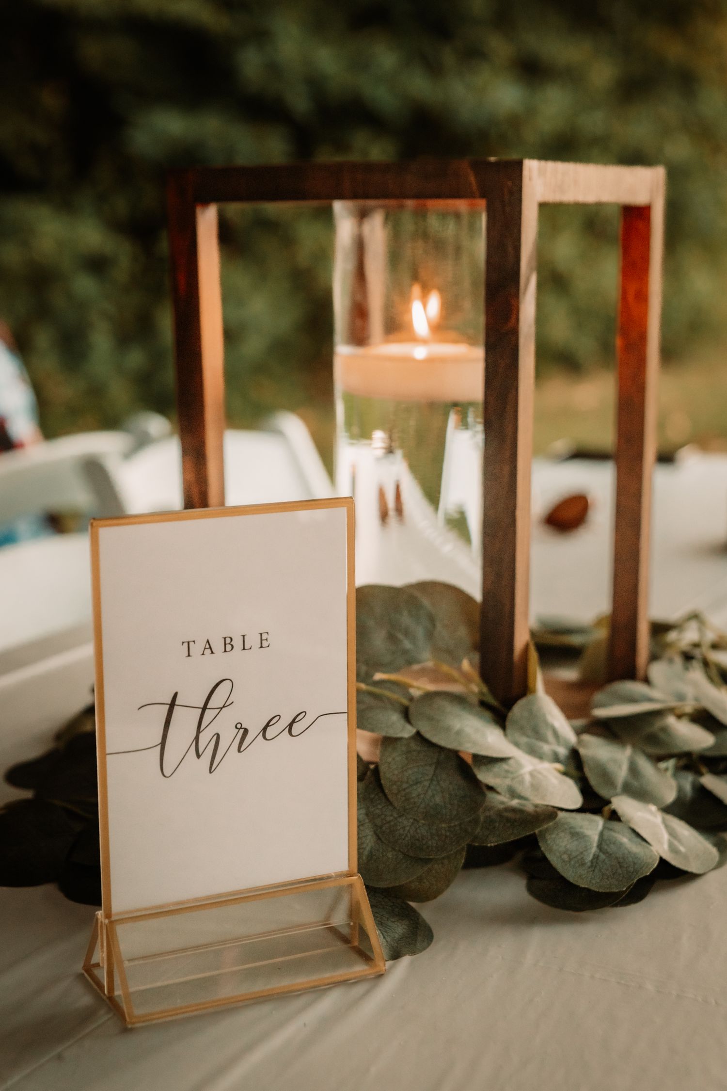 Elegant wedding table number three sign displayed with glass lantern candle holder and eucalyptus leaves on white tablecloth.