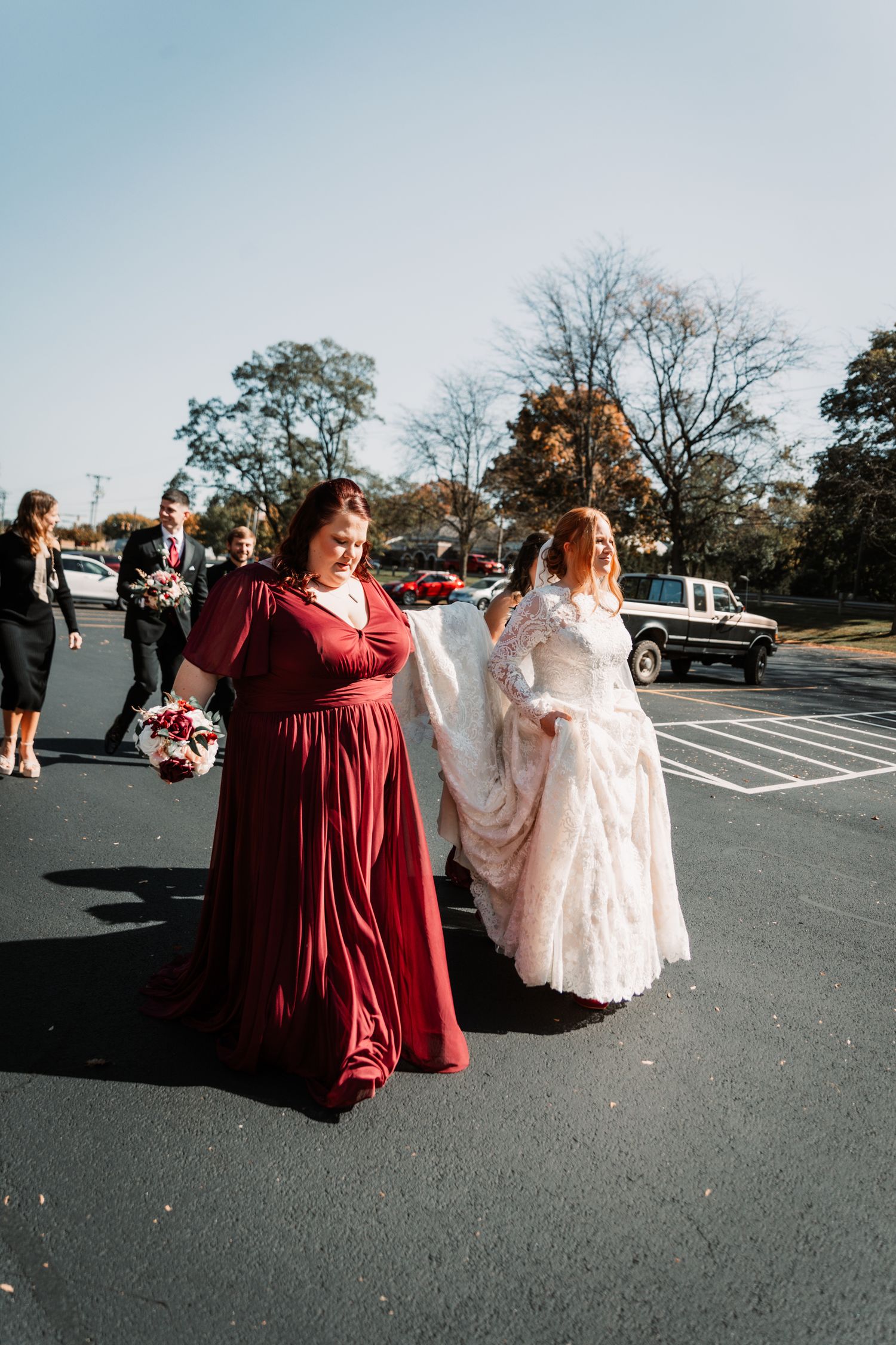 A bridesmaid in burgundy dress and a bride in white lace gown walk together in a parking lot on a sunny day.
