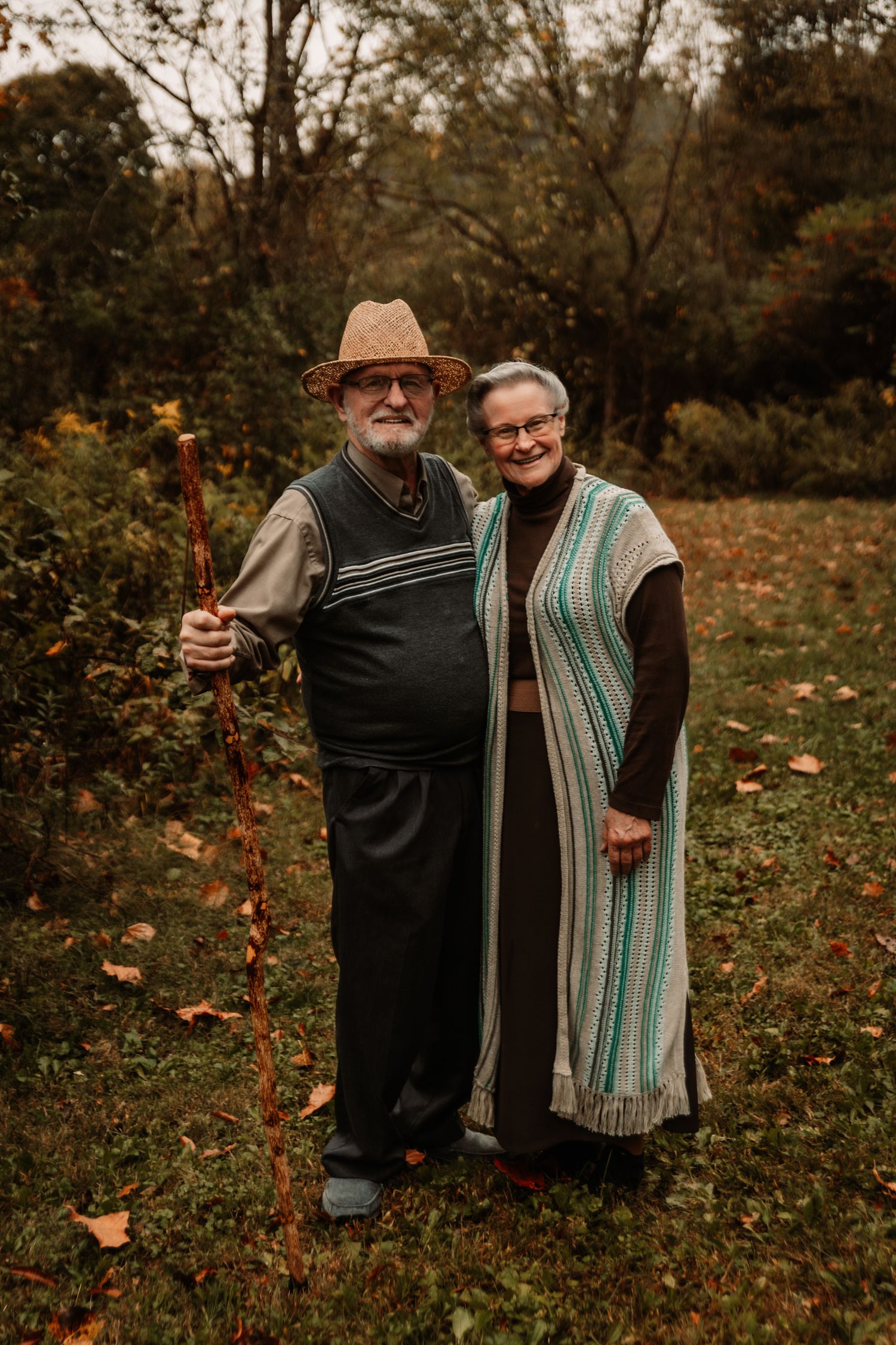 An elderly couple stands together in autumn scenery with fallen leaves, wearing warm clothing and a walking stick.