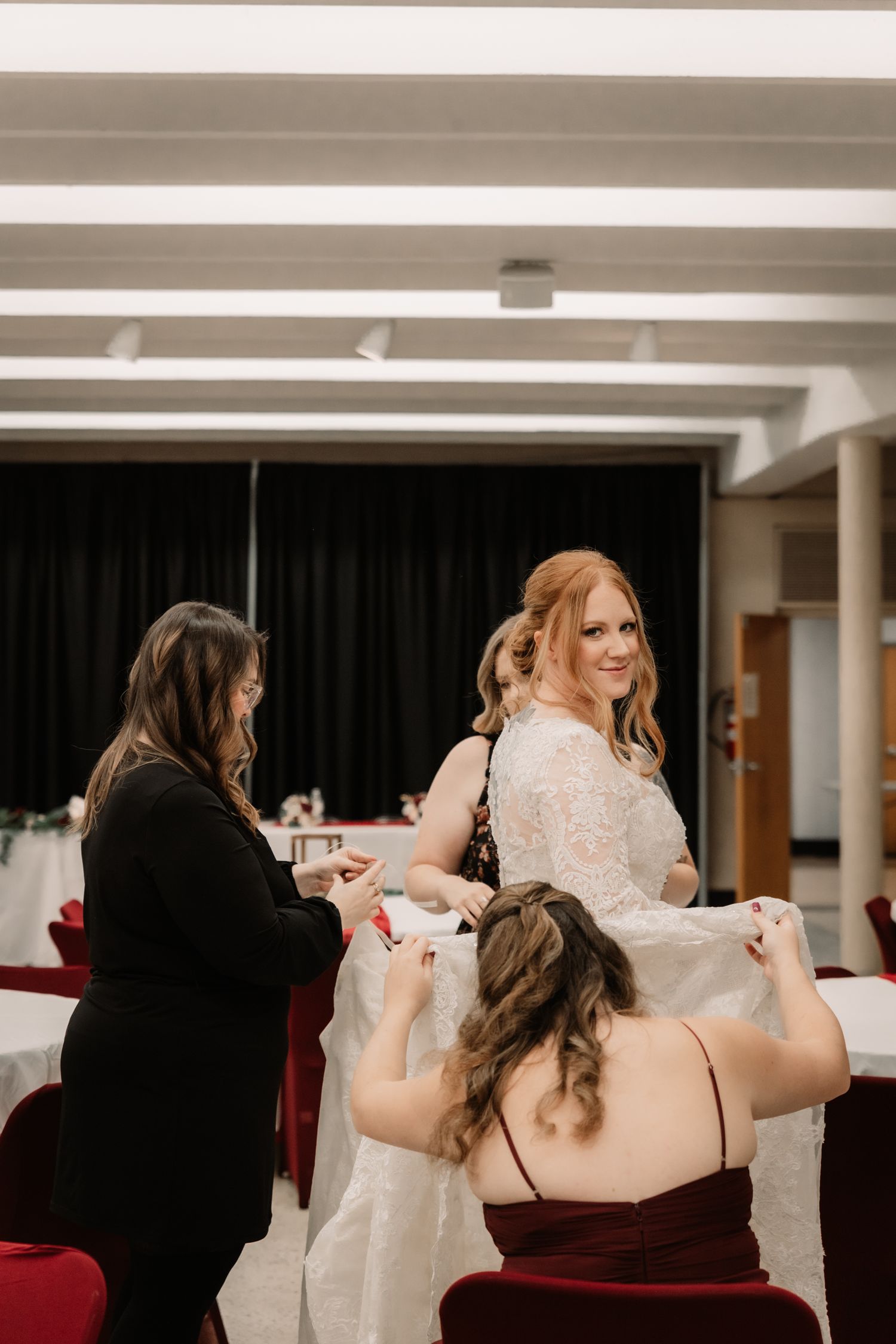 A bride in a long-sleeved lace wedding dress smiles while getting ready with helpers in red chairs.