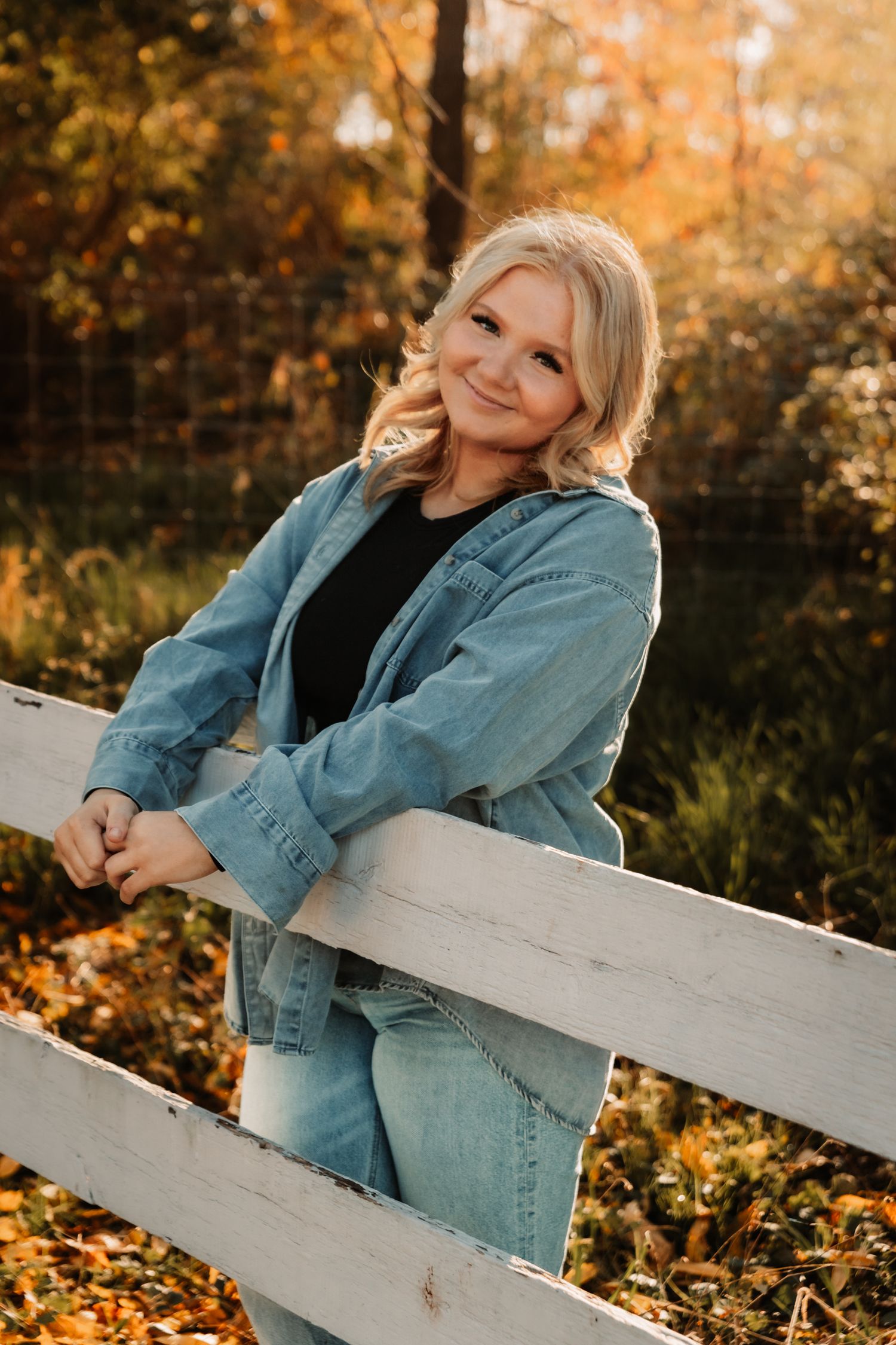Portrait of someone in denim jacket and jeans leaning on a white fence surrounded by autumn foliage at sunset.