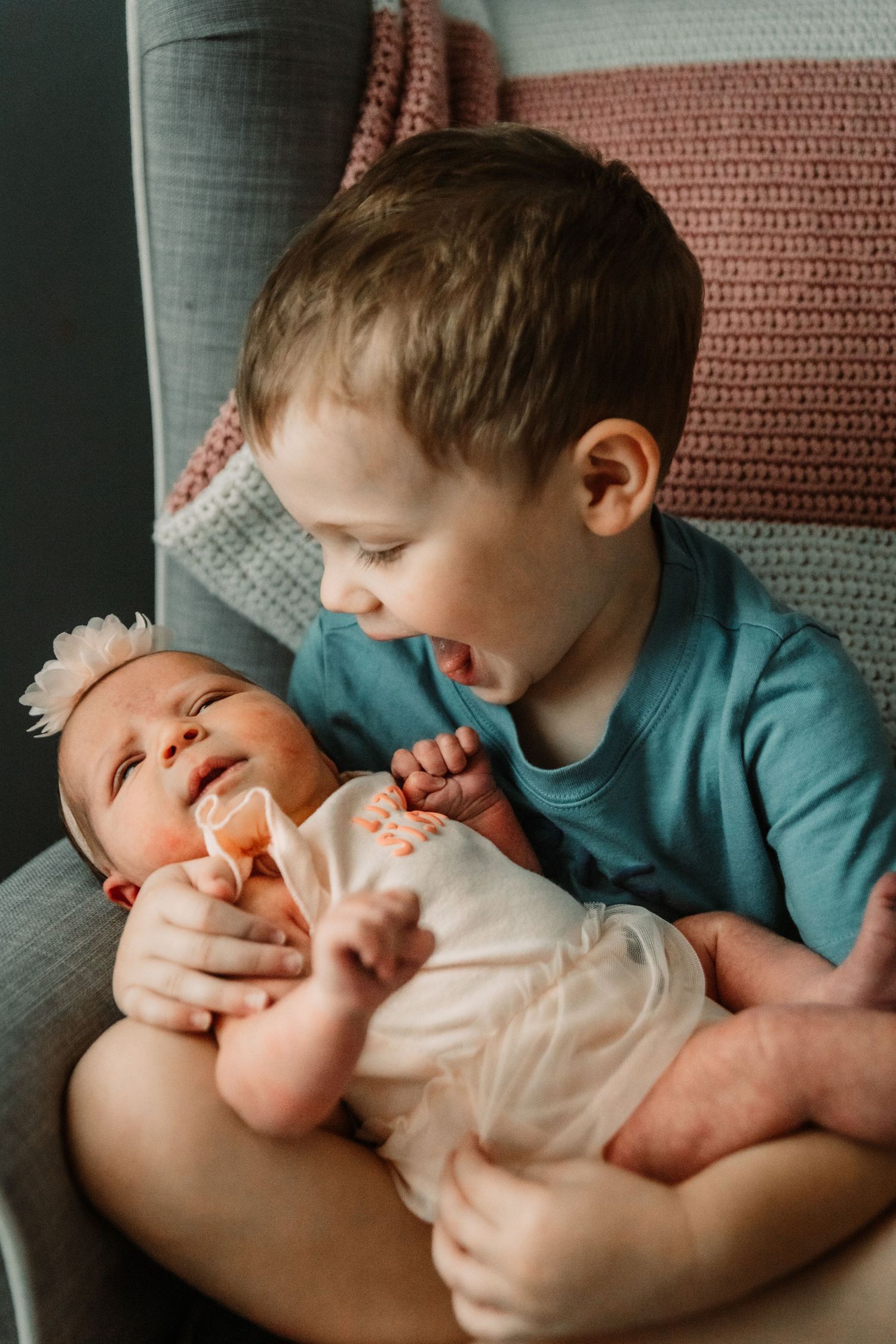 Sweet sibling moment of a child in teal shirt looking adoringly at baby lying on their lap on a gray couch.