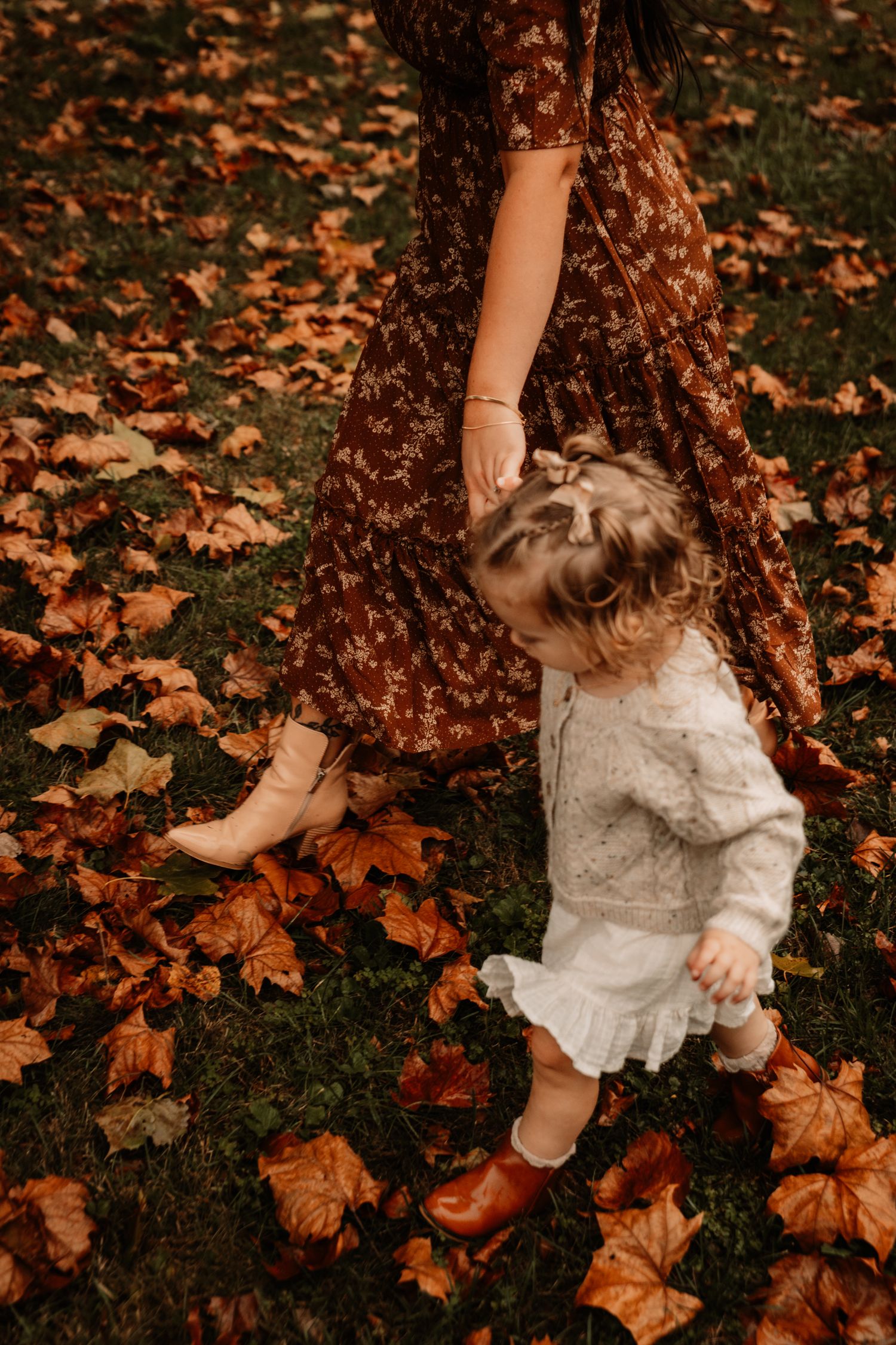 Small child plays among fallen autumn leaves wearing a white dress and brown boots, next to a floral dress.