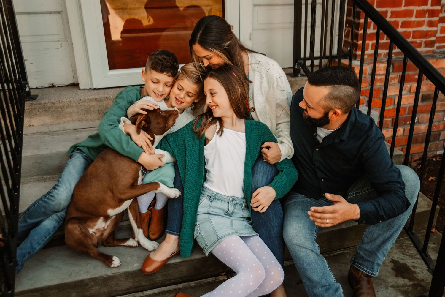 A group of friends in casual clothing laughing together on porch steps with a brown dog.