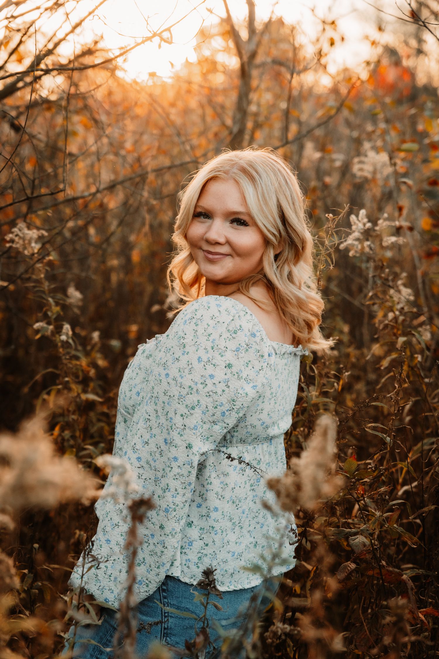 Portrait of someone in a floral top and jeans standing in an autumn field at golden hour with warm backlighting.