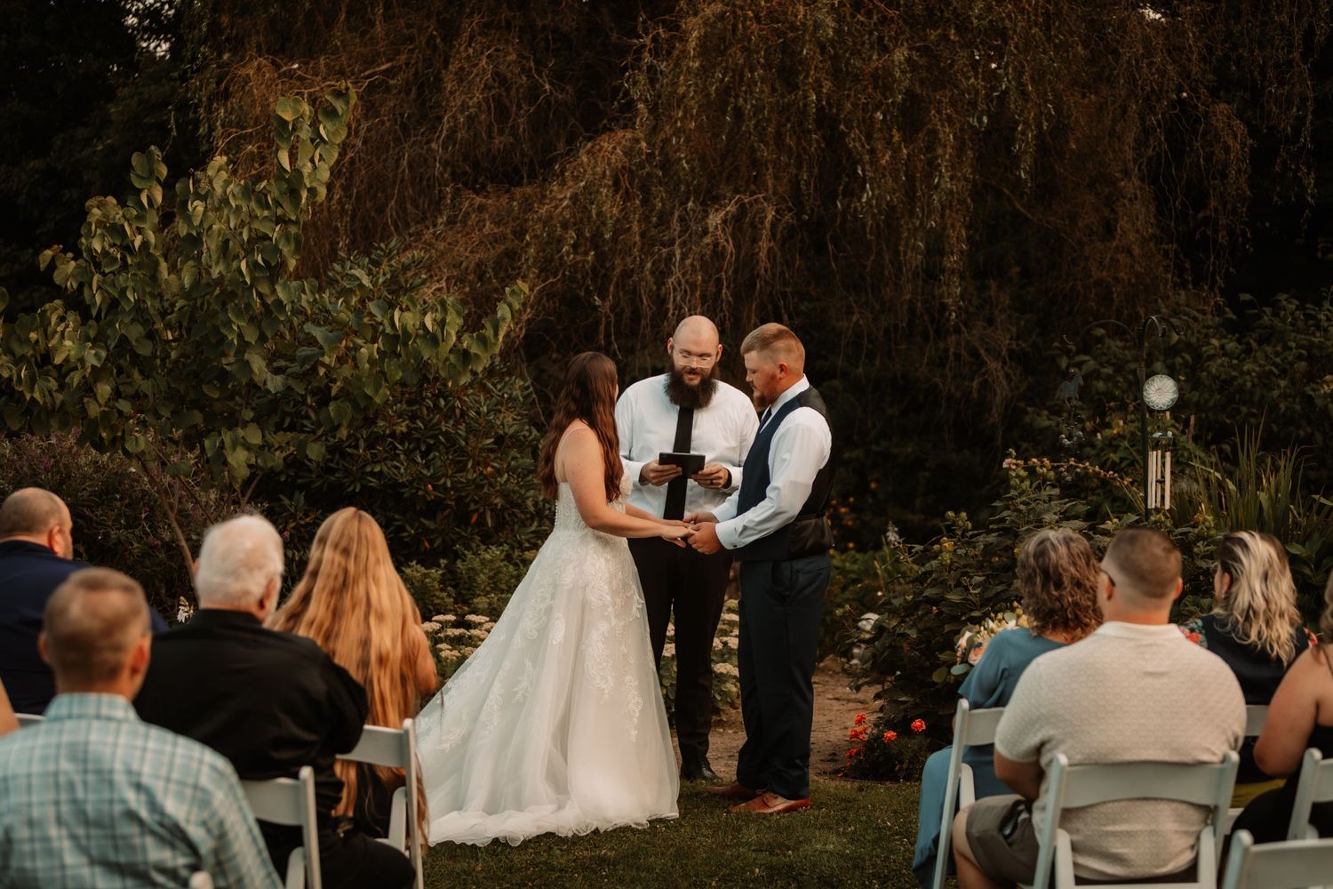 An outdoor wedding ceremony takes places at sunset under a weeping willow tree with guests seated in white chairs.