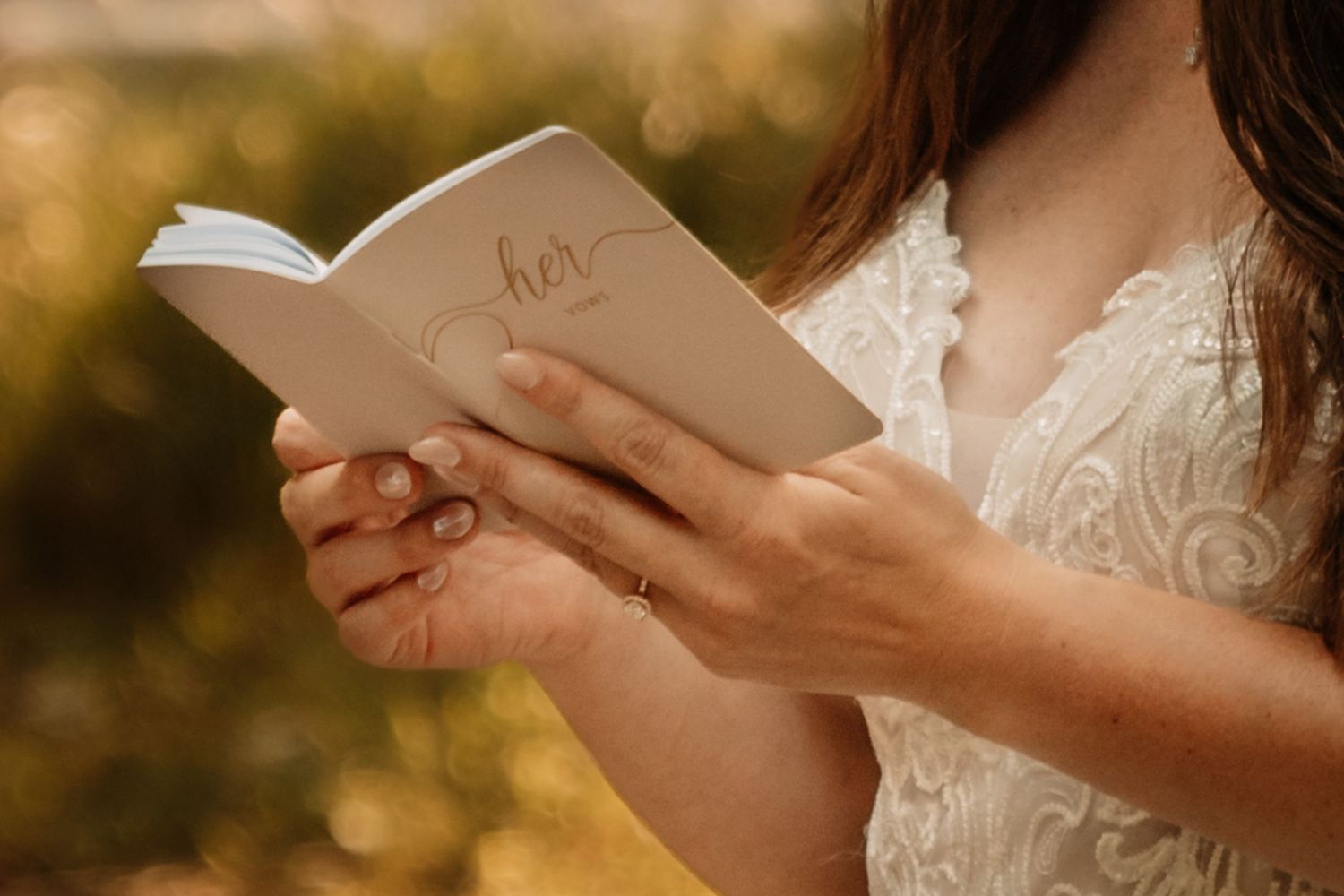 Close-up of hands holding an open book while wearing a white lace dress against a golden bokeh background.