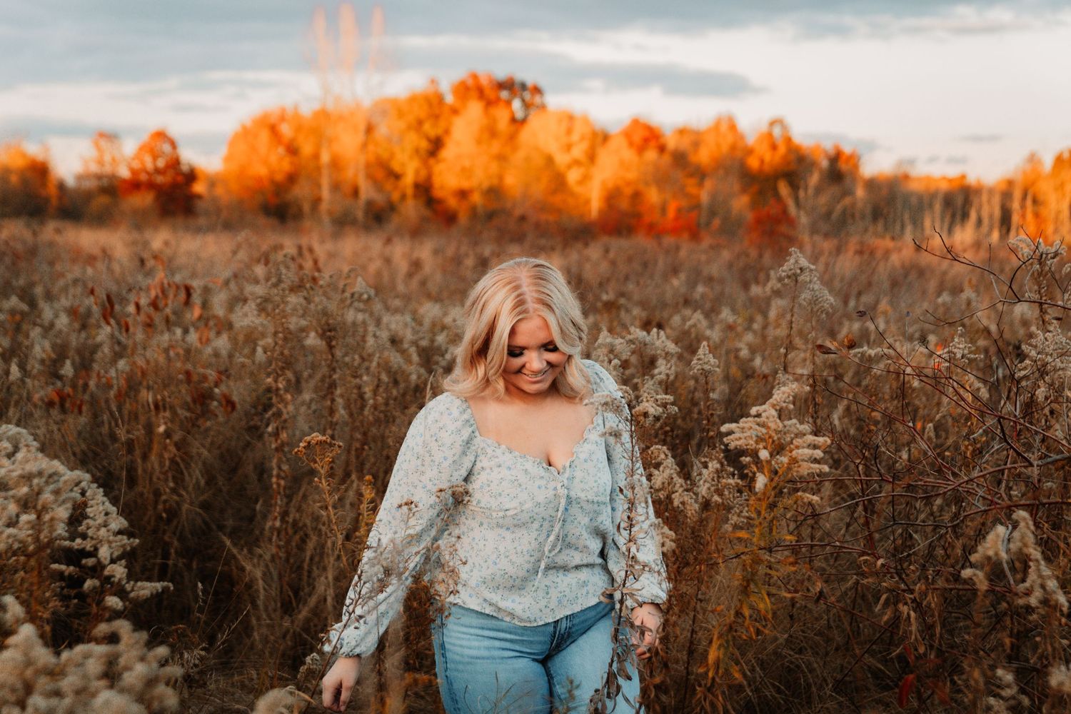 A person in a light sweater walks through a golden autumn field at sunset with warm glowing light on dried grasses.