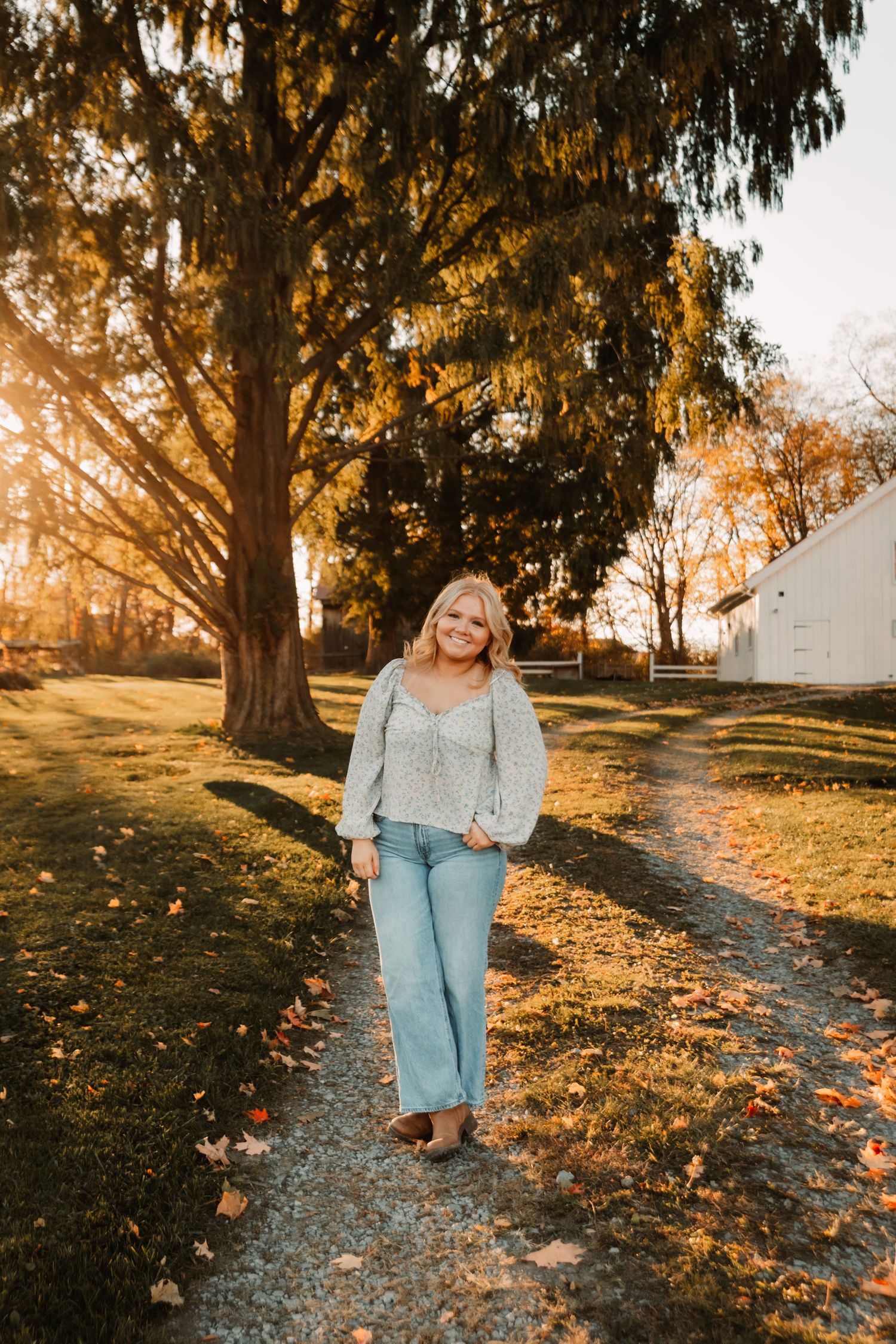A person walks along a gravel path at sunset, surrounded by autumn trees and fallen leaves in golden light.