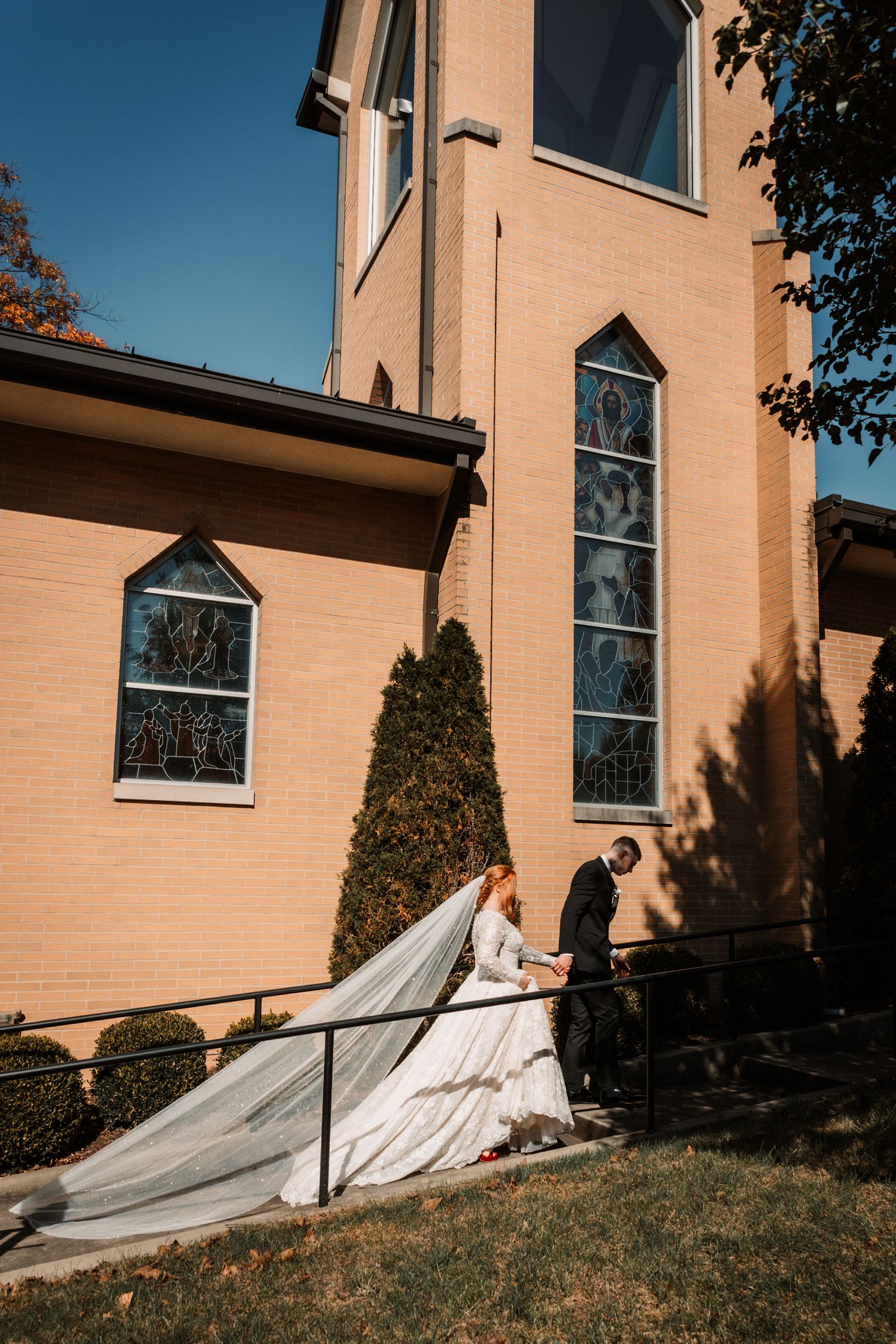 An elegant couple in wedding attire stands outside a tan church with tall stained glass windows on a sunny day.