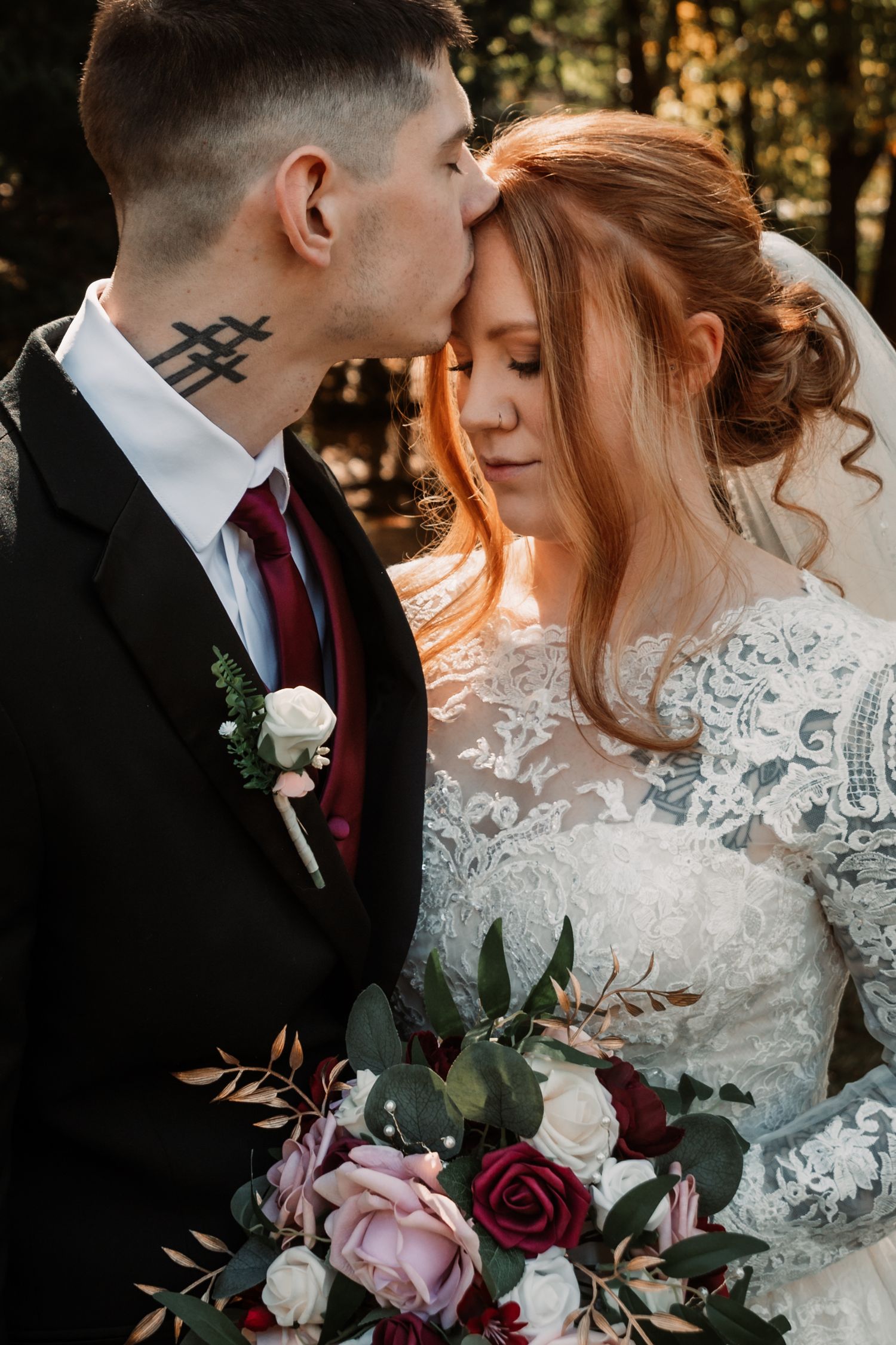 Intimate wedding portrait shows a couple with their foreheads touching, bride holding burgundy and pink bouquet in lace gown.