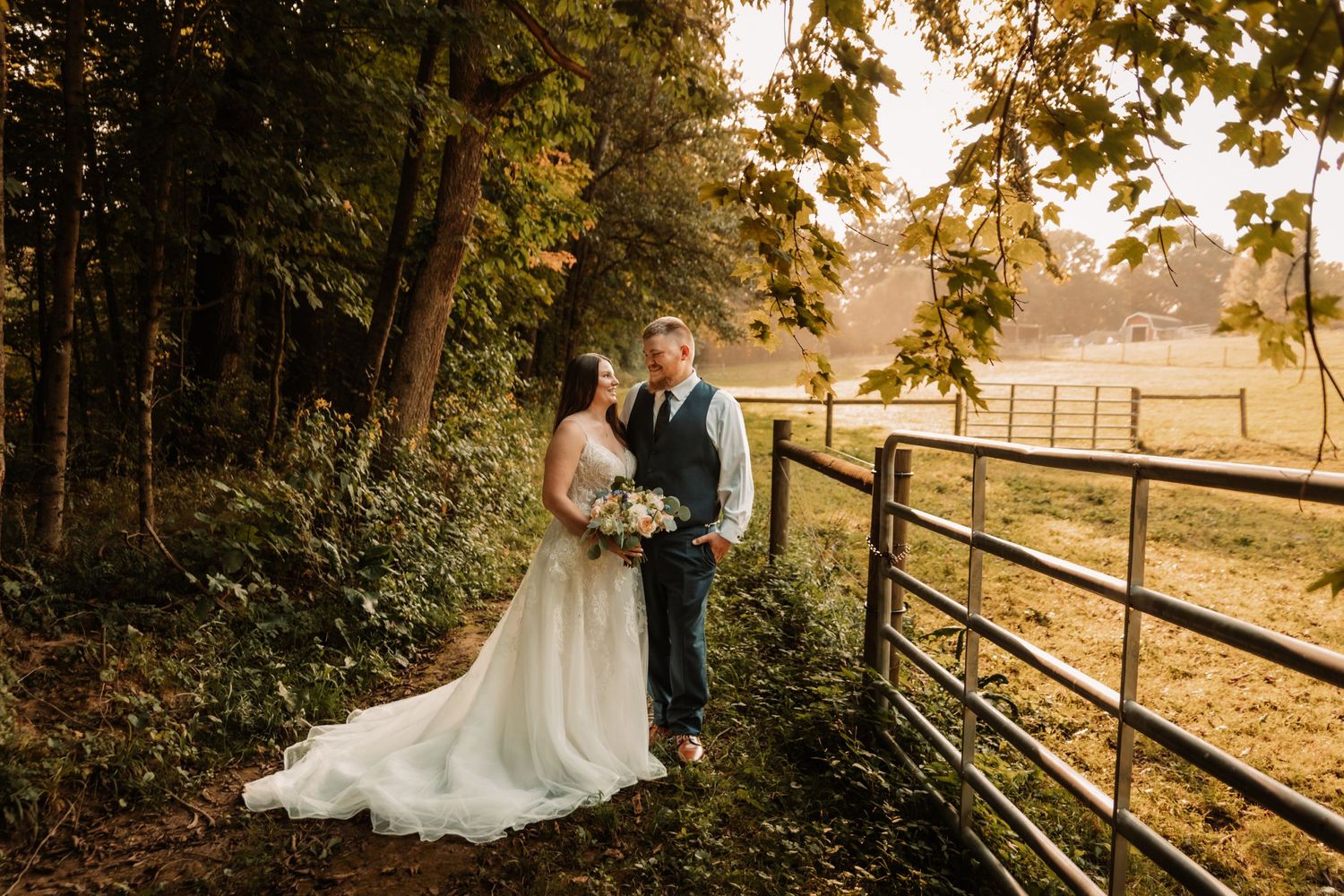 A romantic autumn wedding photo at sunset by a rustic farm gate with a warm golden glow through the trees.