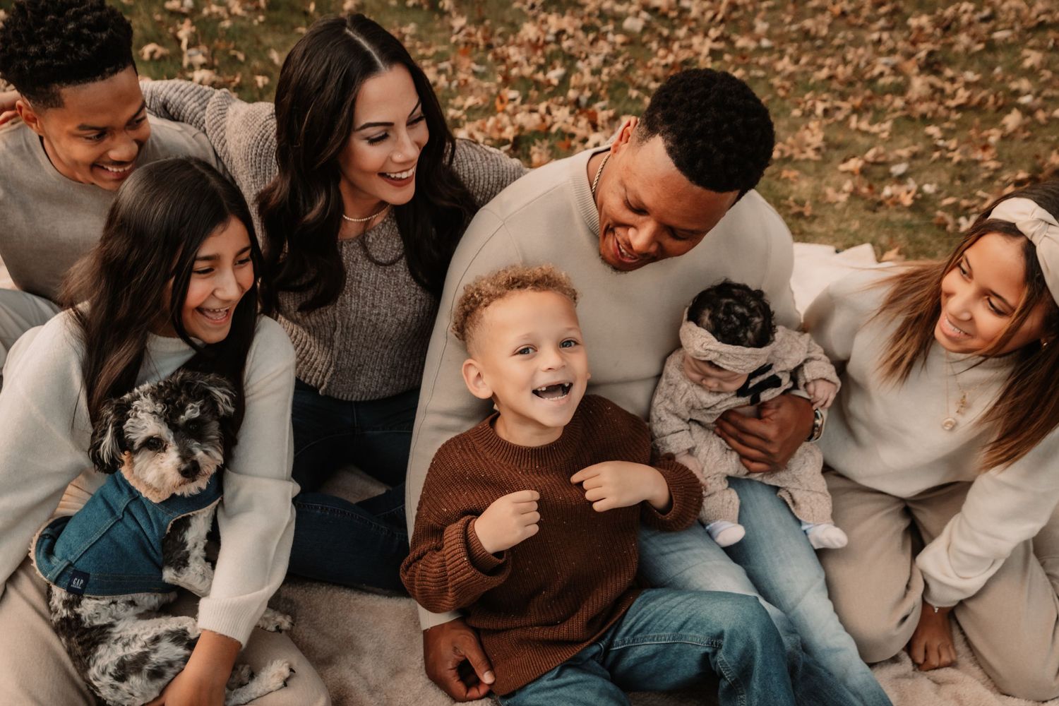 A group in sweaters sits together outdoors on fallen autumn leaves with their pets while sharing a joyful moment.