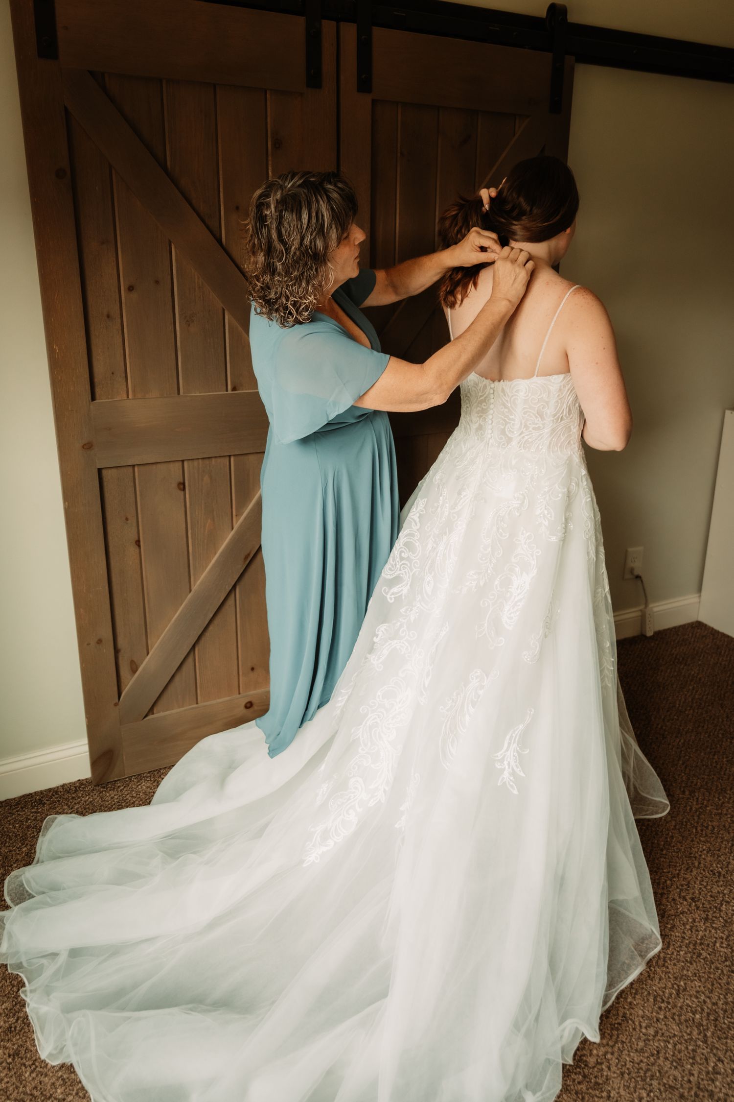 Person in turquoise dress helps adjust a stunning white wedding gown with long train against rustic barn doors.