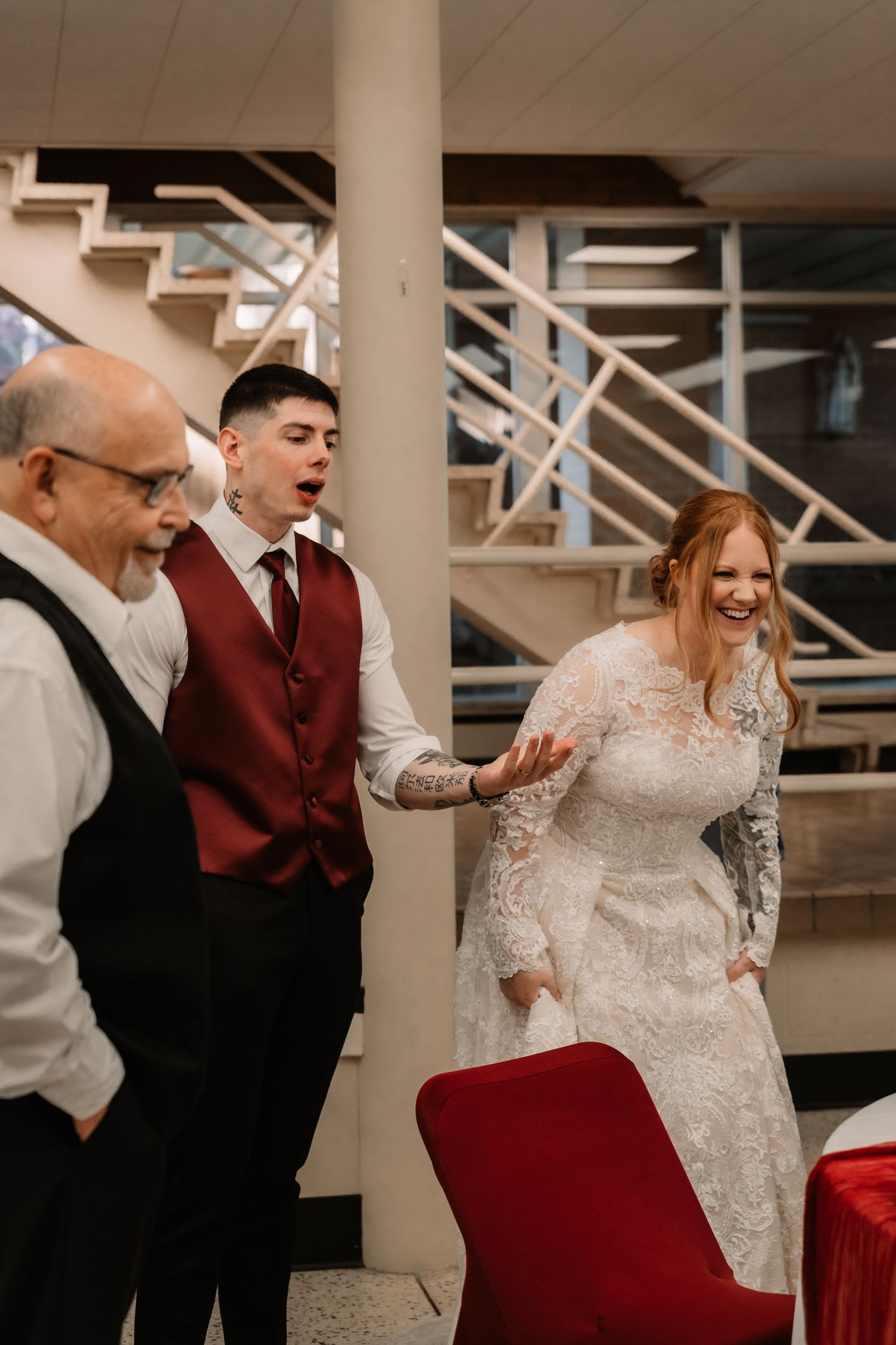A candid reception moment shows guests in red vests and a bride in an elegant long-sleeved lace gown laughing together.