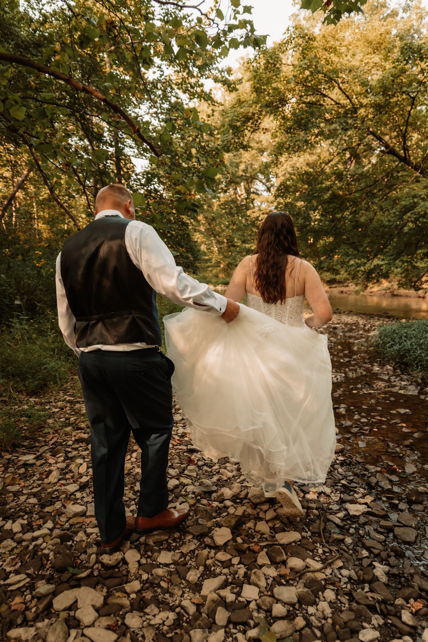 A romantic wedding photo of a couple walking along a rocky riverbed surrounded by autumn trees.