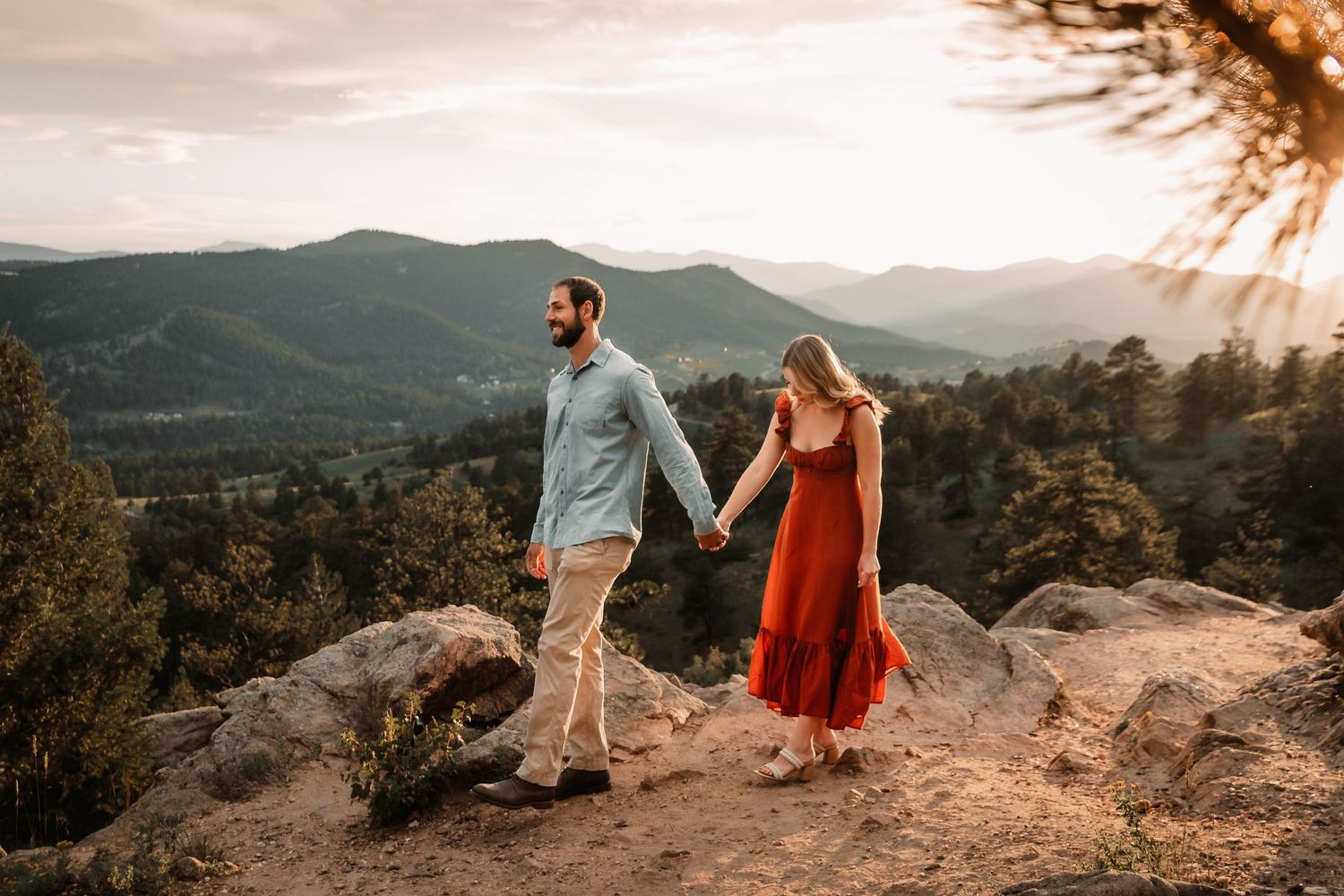 Two people holding hands while standing on a mountain overlook during golden sunset in forested hills.