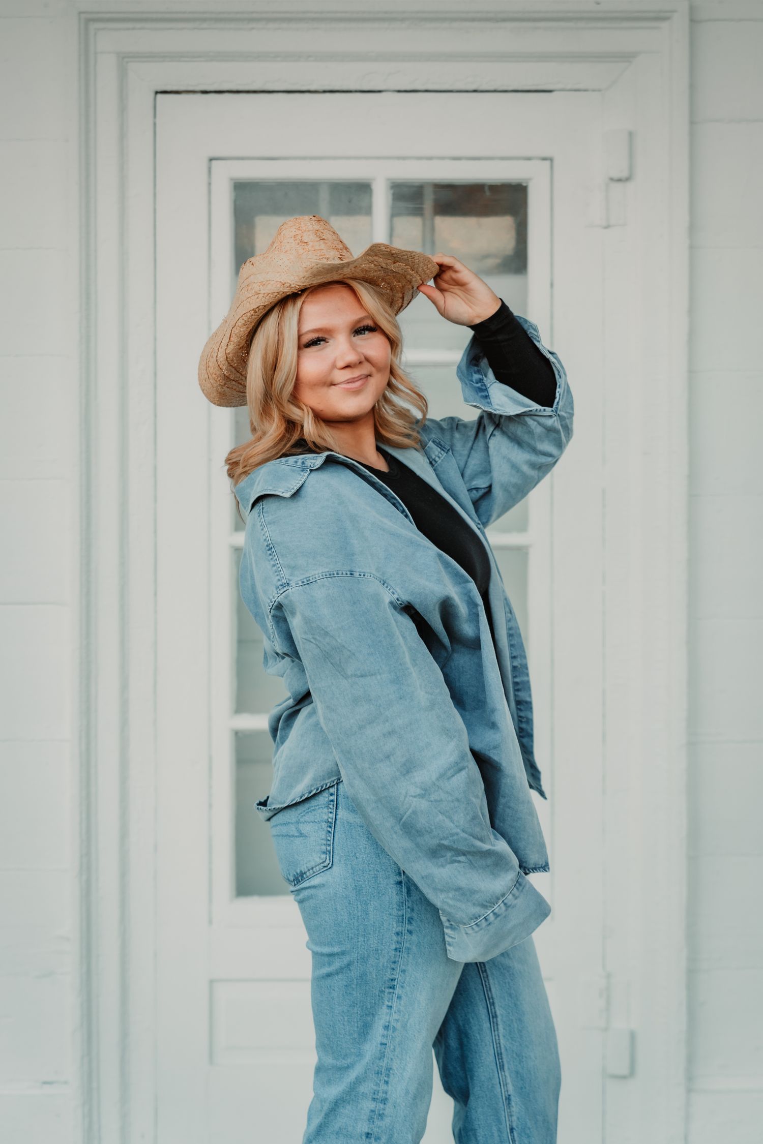 A person wearing a denim jacket and straw hat poses against a white door in a casual, country-inspired style.