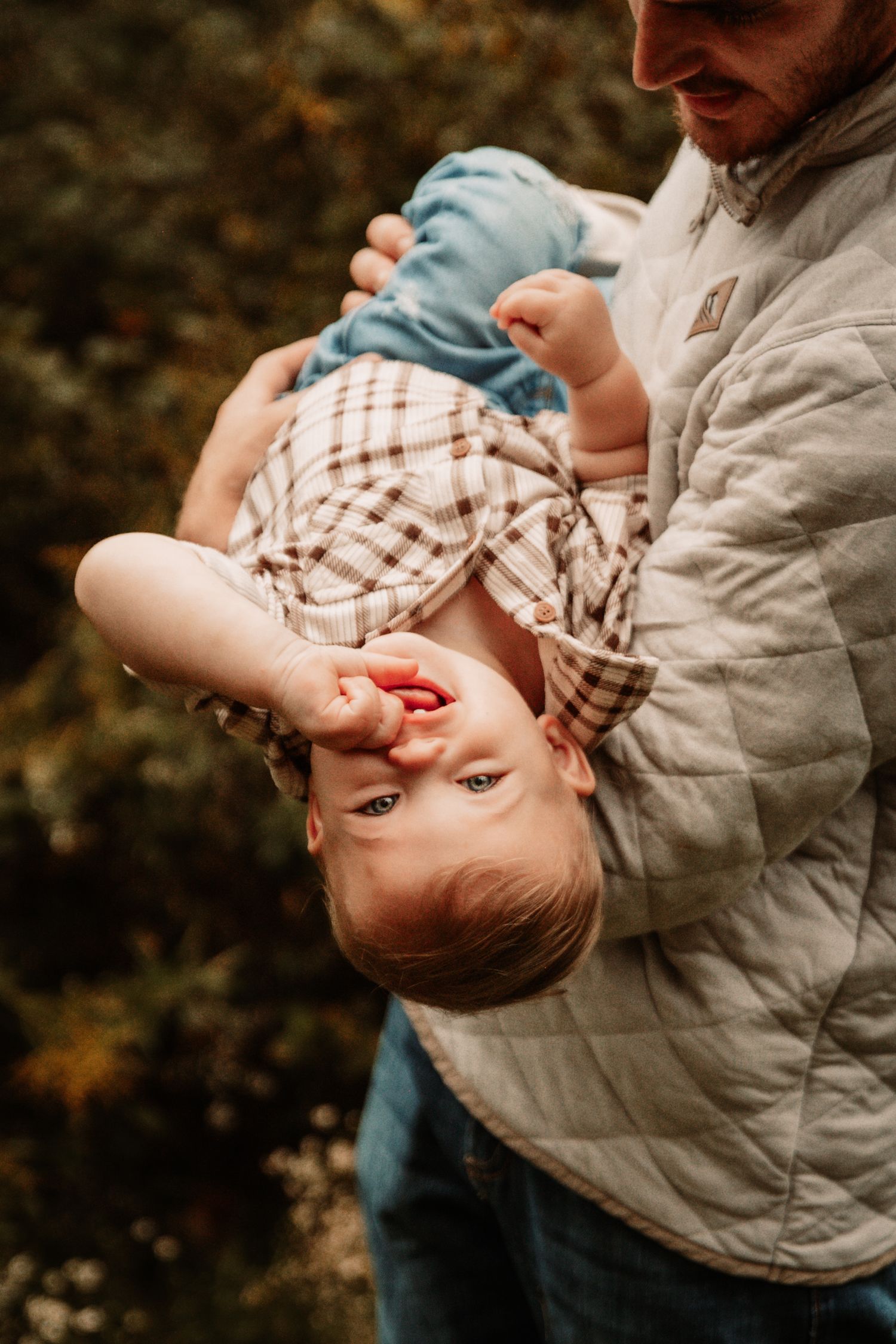 A young child in a plaid shirt being playfully held upside down by someone wearing a quilted jacket outdoors.
