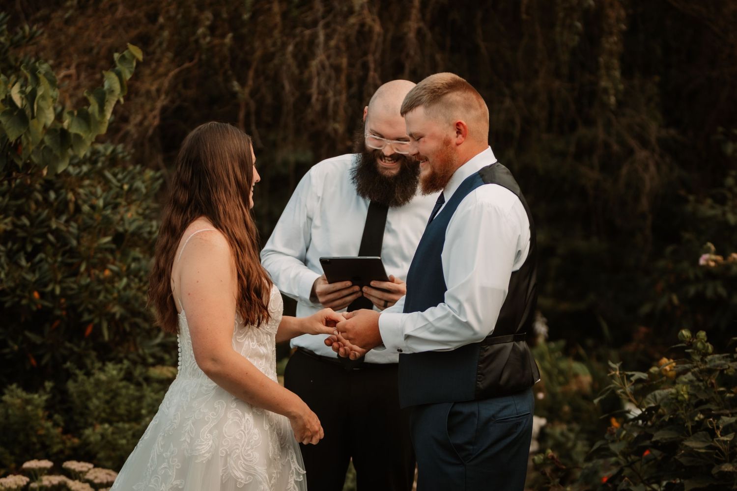 Outdoor wedding ceremony taking place in a lush garden setting at sunset with three people in formal attire.