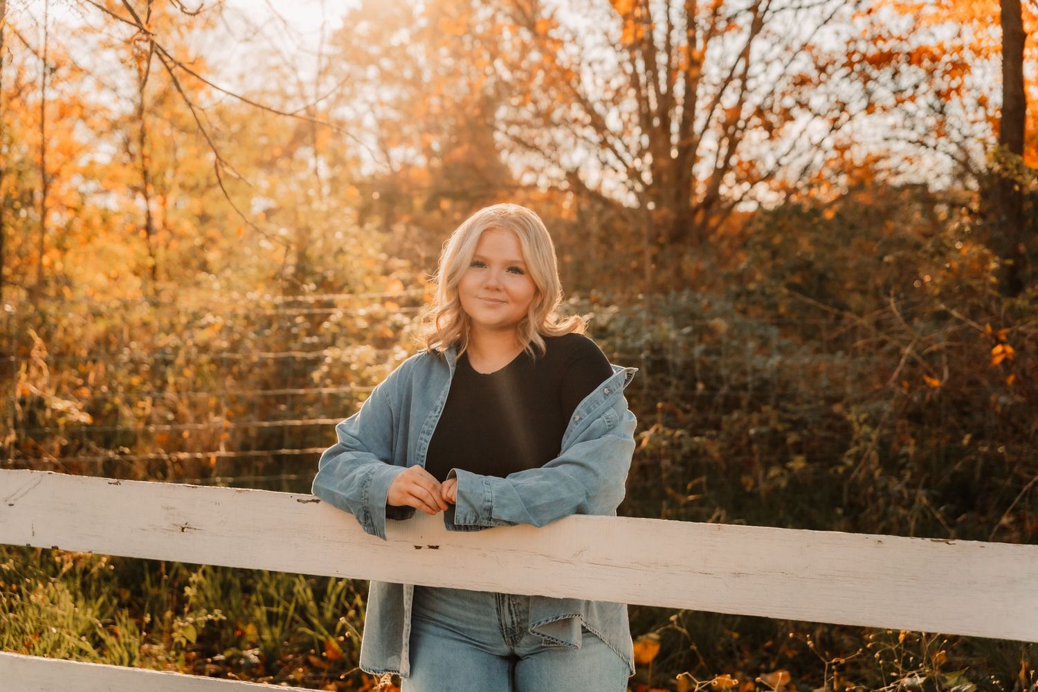 Someone in a denim jacket leans against a white wooden fence during golden hour in an autumn setting.