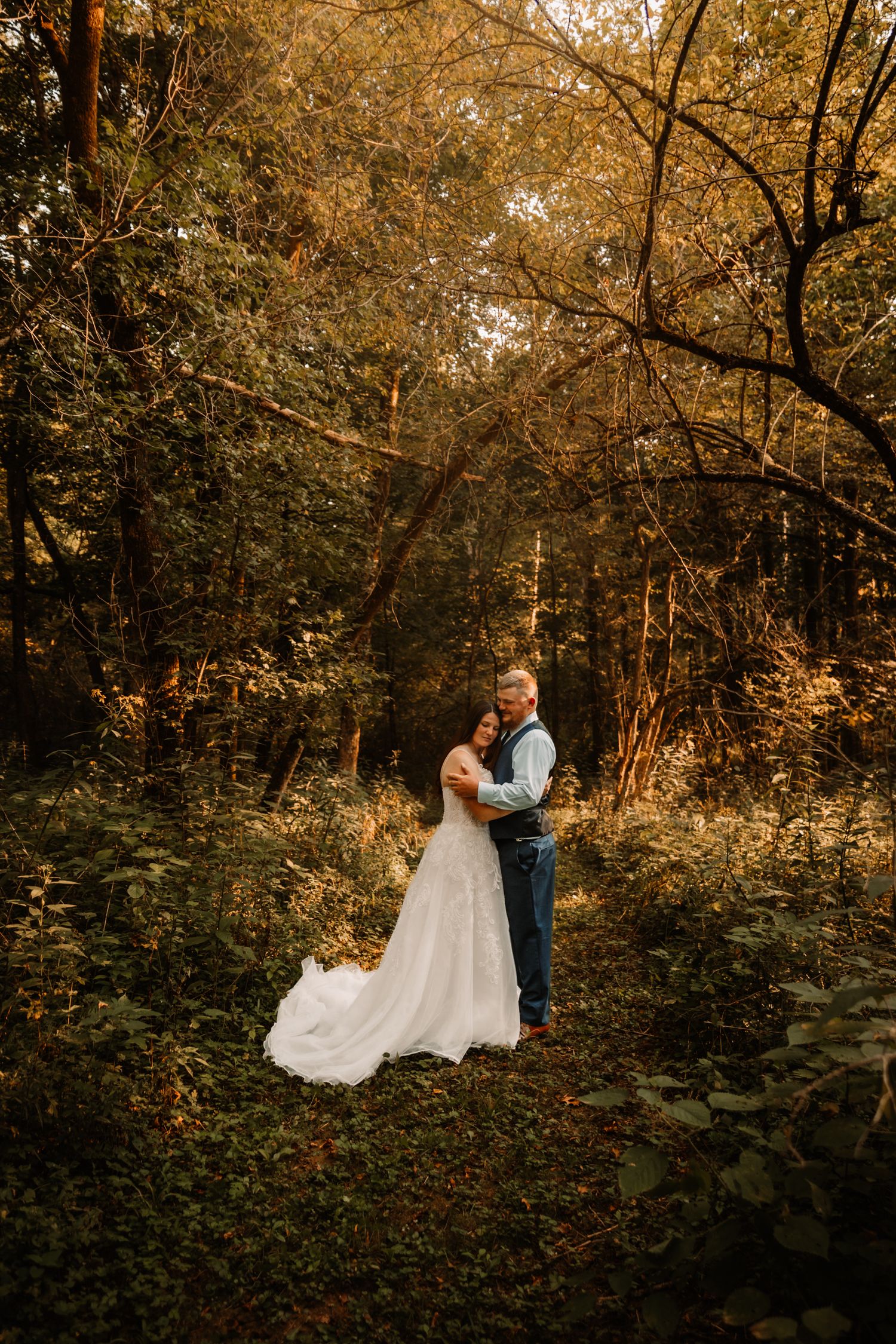 A romantic wedding couple embraces in a golden-lit autumn forest with sunlight filtering through the trees.
