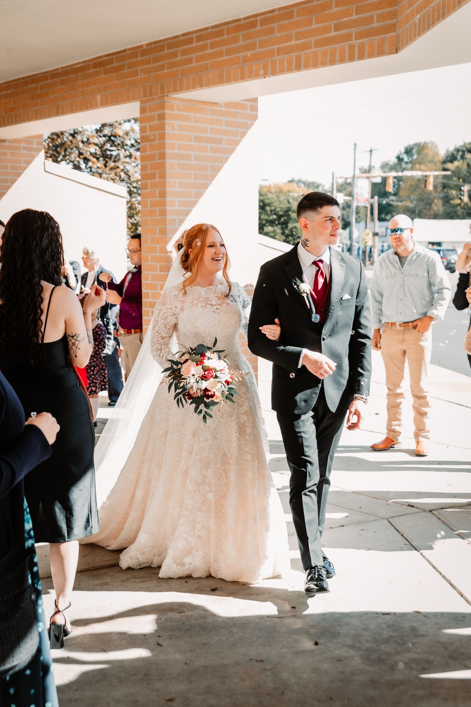 Newlyweds walk down a sunlit brick hallway as wedding guests look on during a joyful celebration.