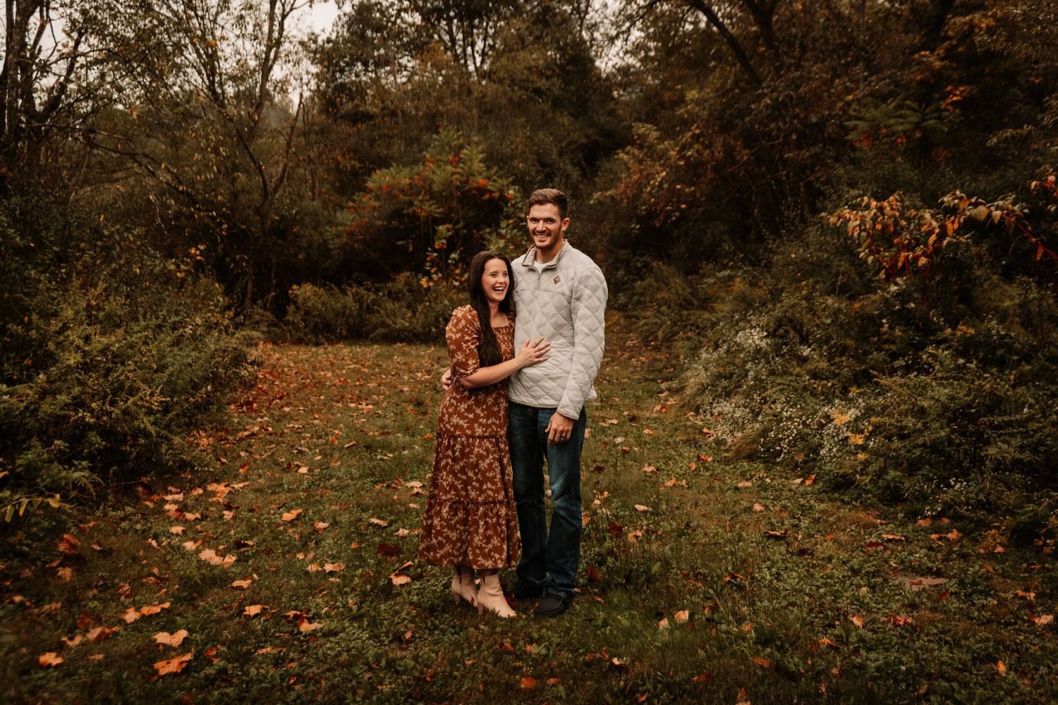 A couple stands close together in casual attire against a backdrop of fall foliage and leaves.