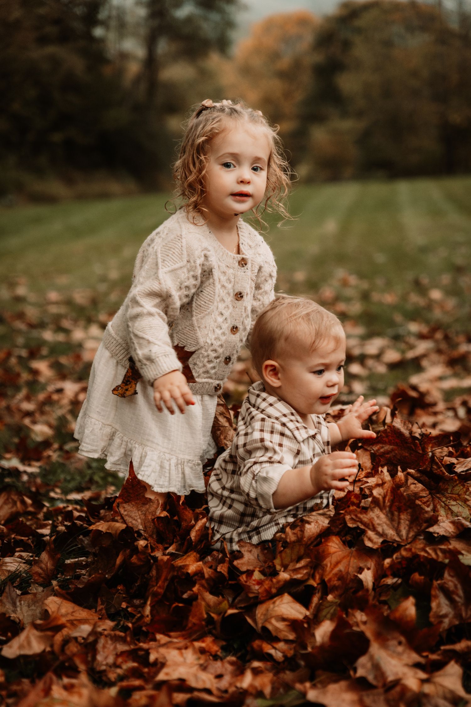 Two small children in vintage-style clothing playing together in a pile of autumn leaves on a fall day.