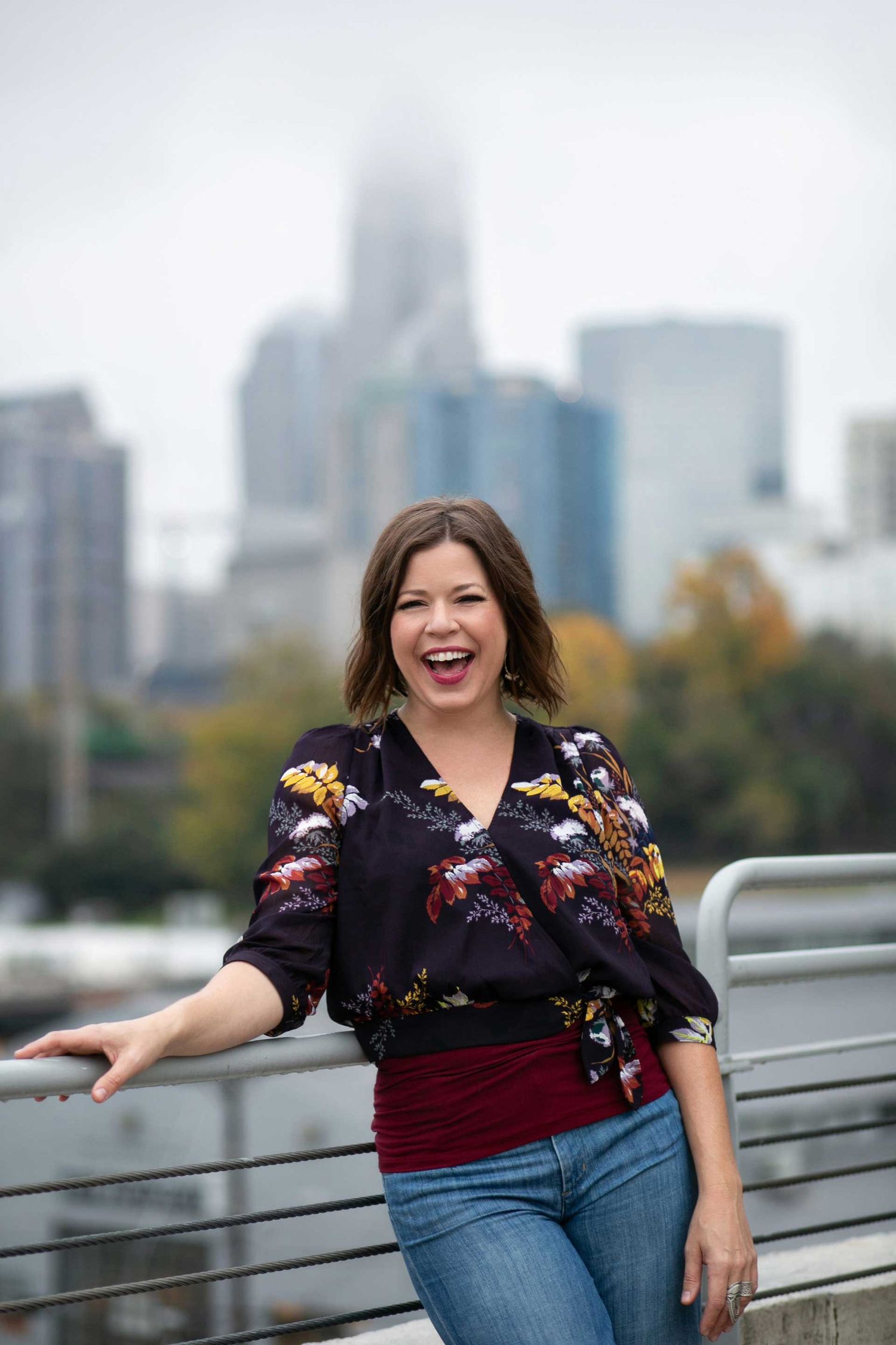 Cheerful person wearing floral navy blouse and burgundy waistband poses by railing with city skyline in background.