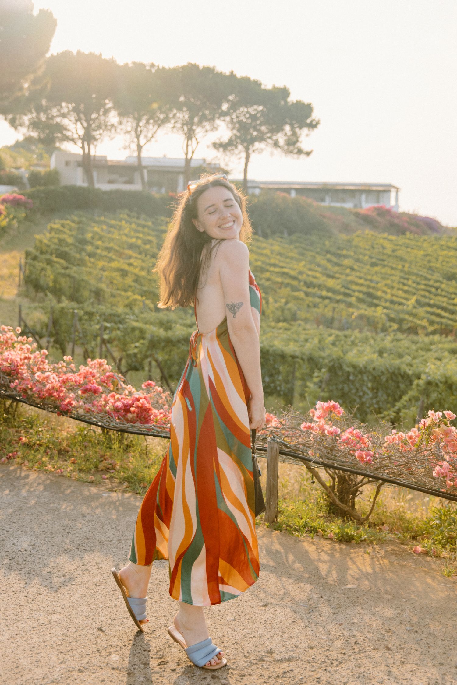 Smiling woman in colorful striped maxi dress poses at golden hour near vineyard with pink flowers.