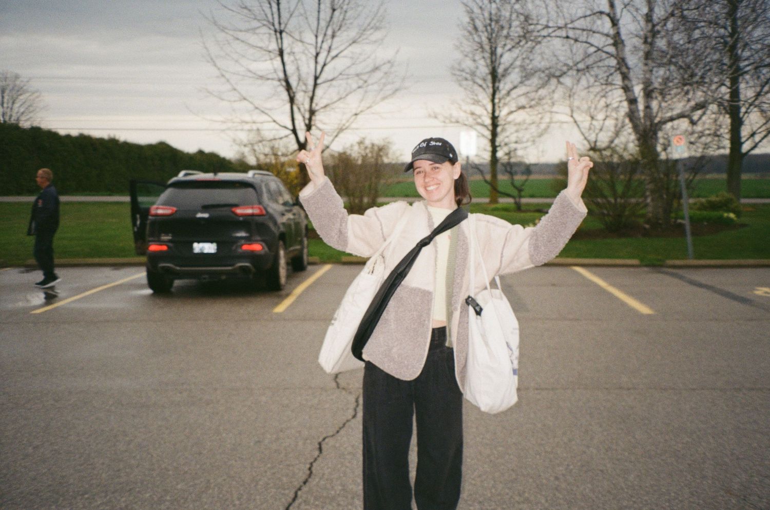 Person in cream jacket and cap poses with arms raised in parking lot at dusk.
