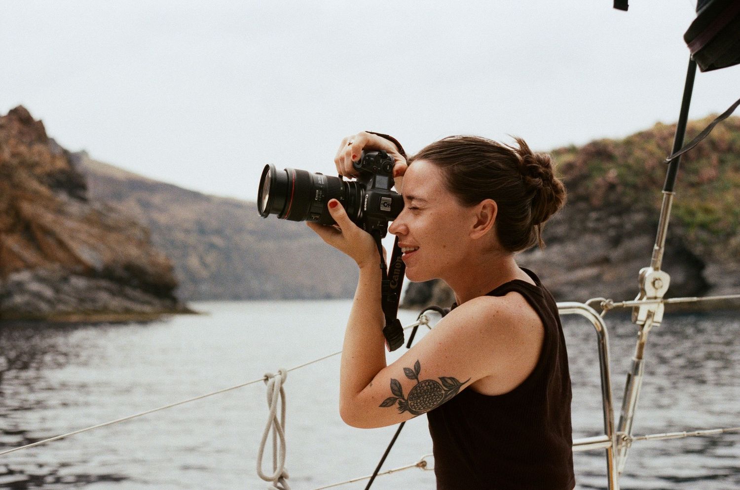 Tattooed woman photographing scenic rocky coastline from sailboat deck with DSLR camera.