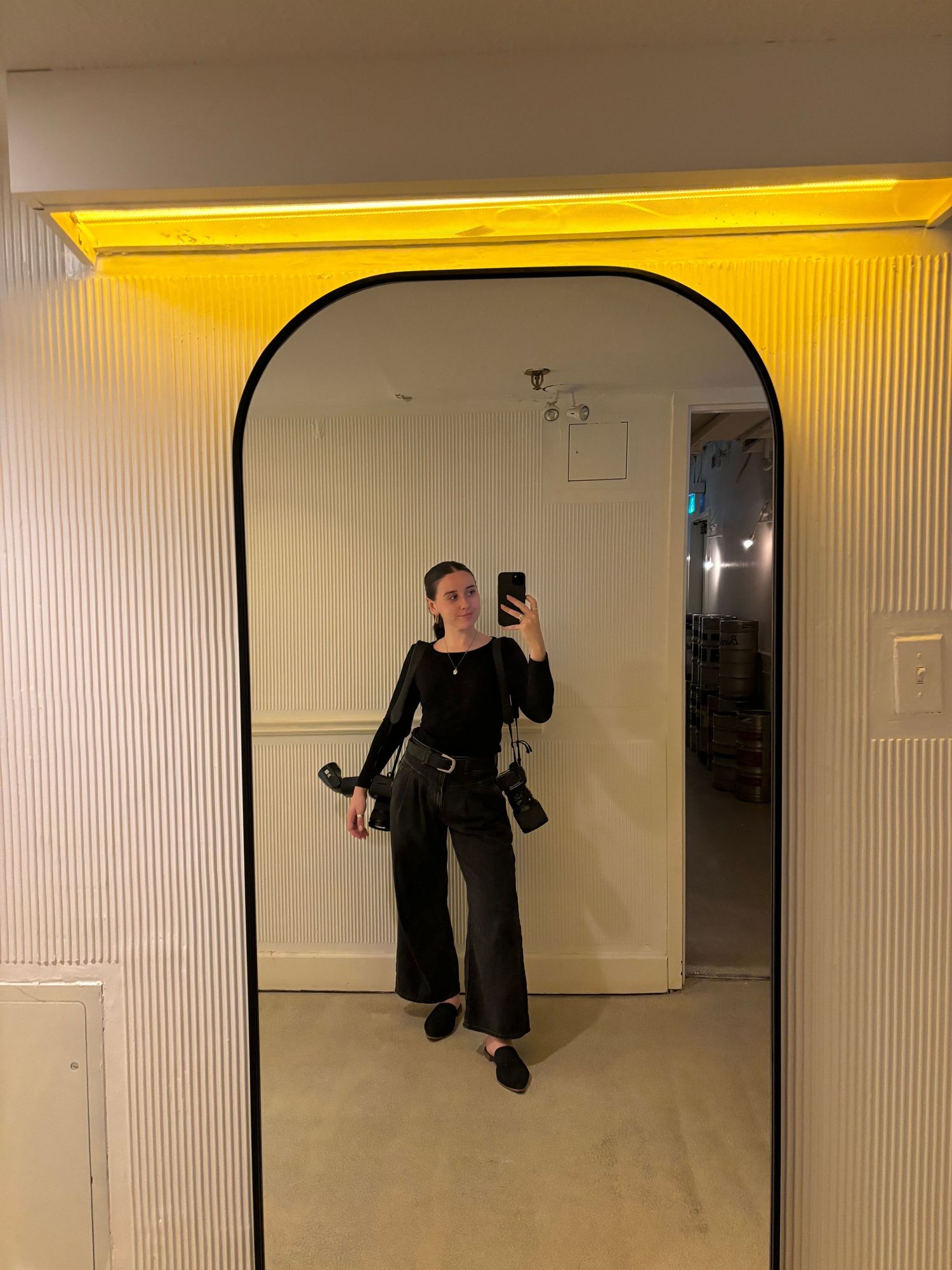 Woman in all black outfit taking mirror selfie with yellow neon light overhead.
