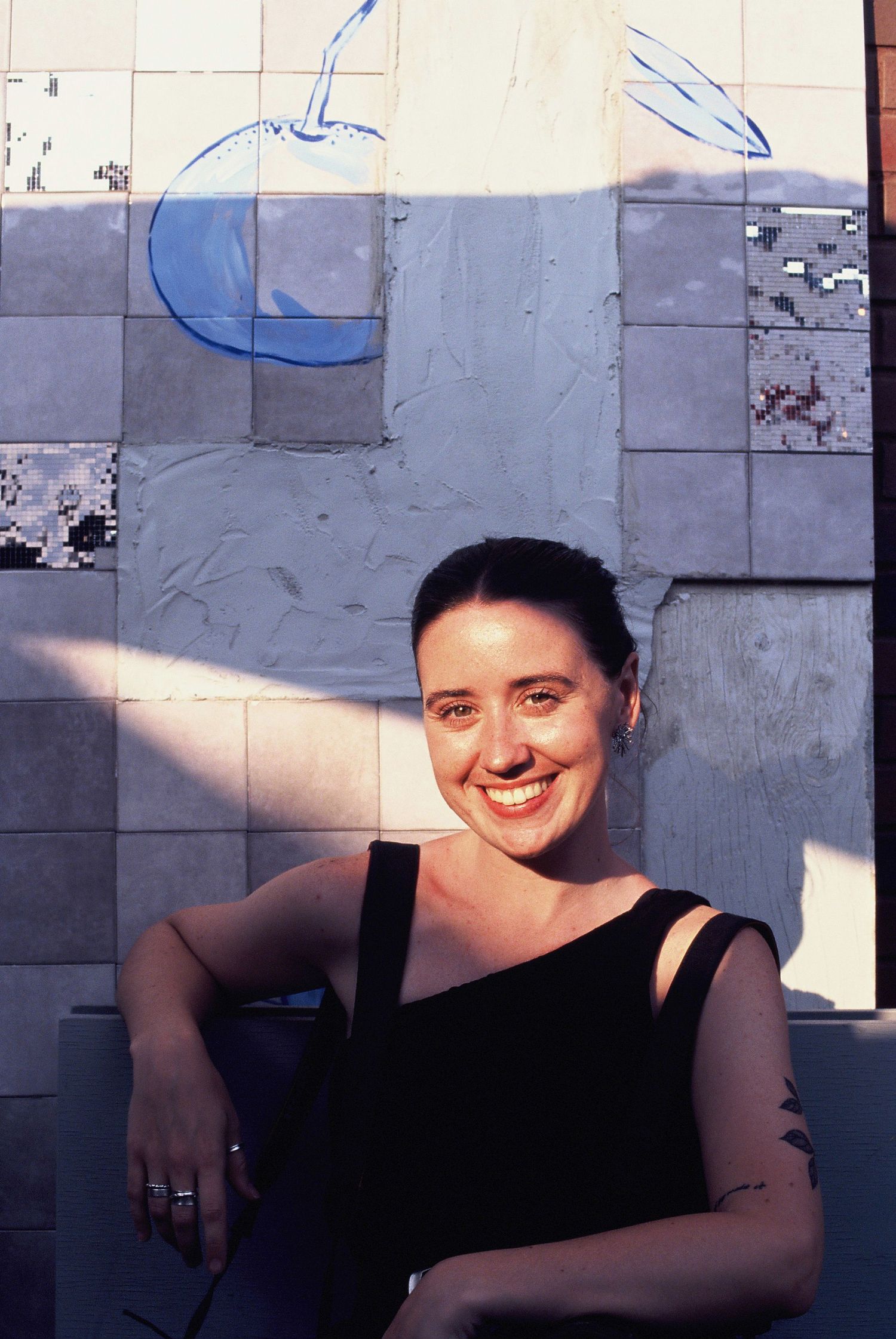 Smiling woman with short dark hair sitting against tiled wall with blue bird mural.