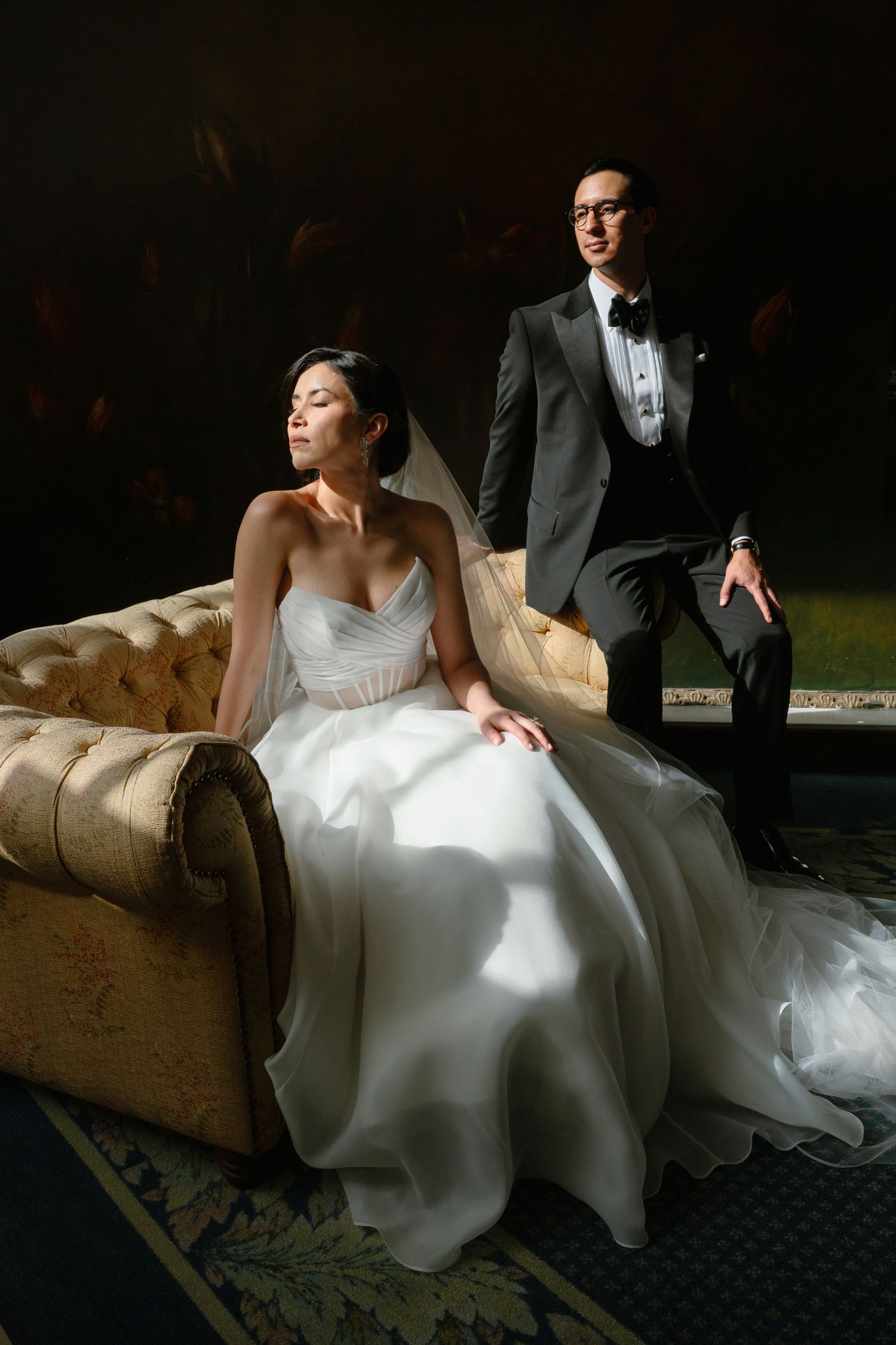 Bride in white gown sits on vintage chaise while groom in gray tuxedo stands behind her in elegant wedding portrait.